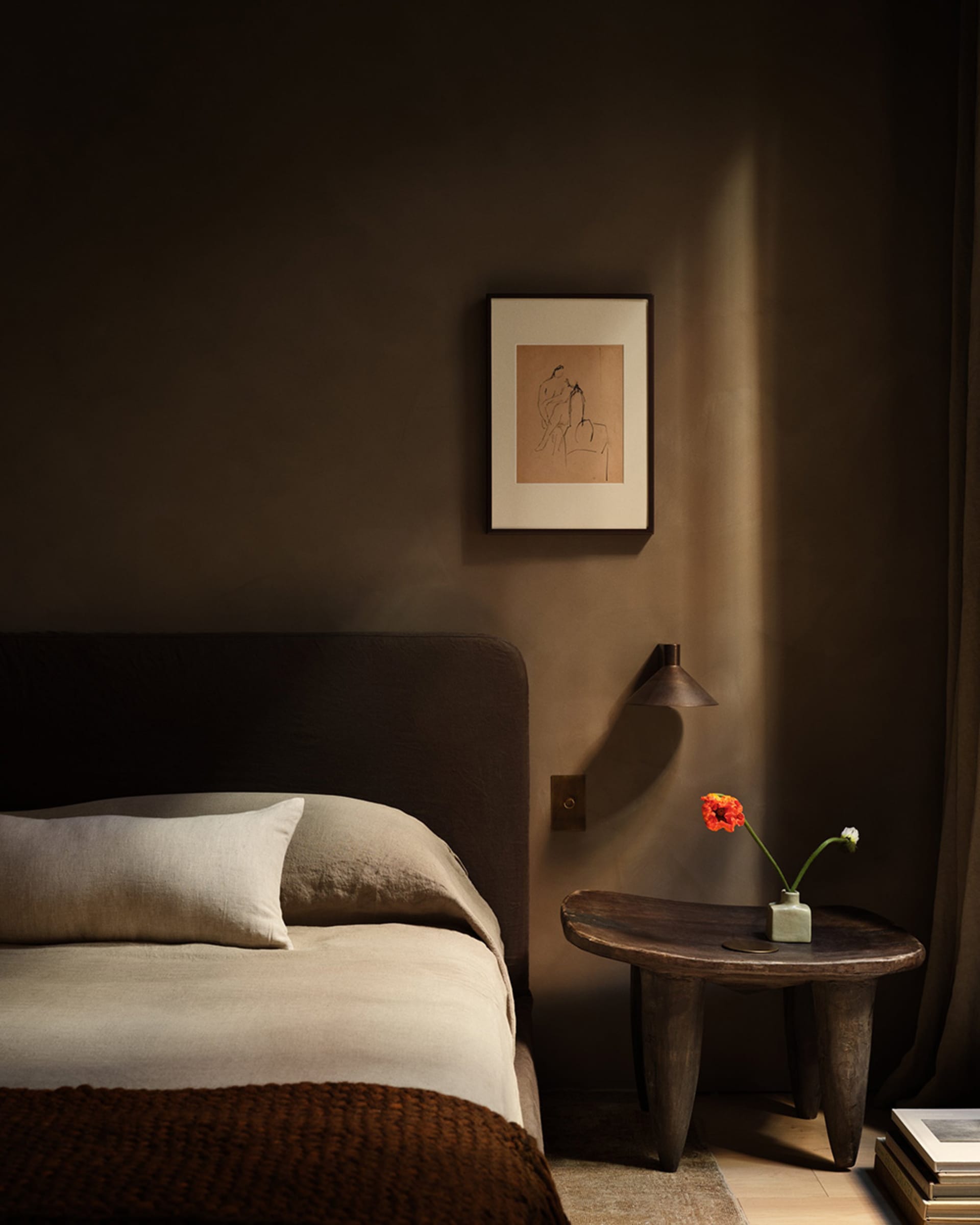 A minimalist bedroom featuring a bed with a beige comforter and white pillows, a wooden bedside table with a vase of flowers,
