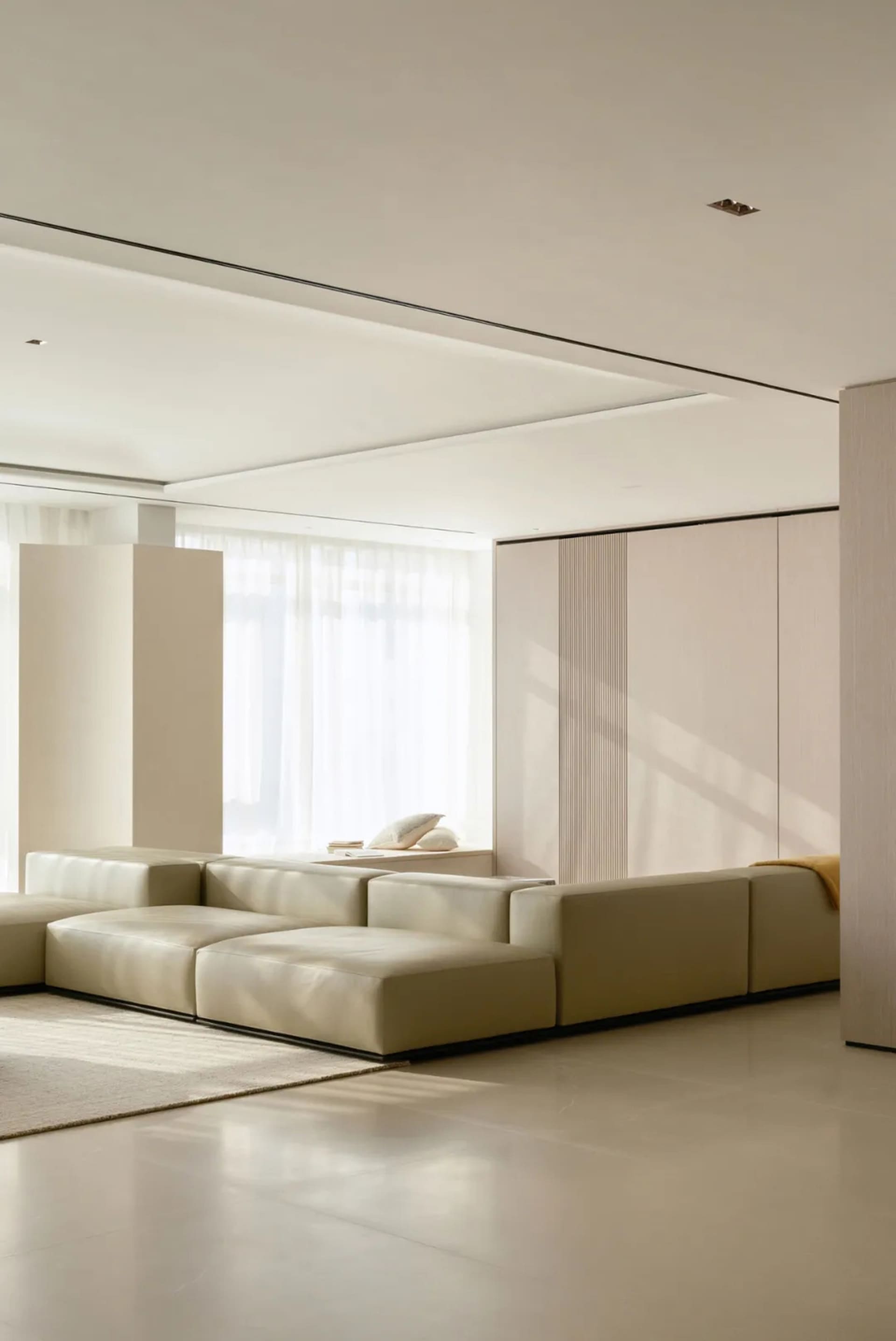 A modern minimalist living room with a large beige sectional sofa and a light-colored rug, featuring soft, neutral tones and