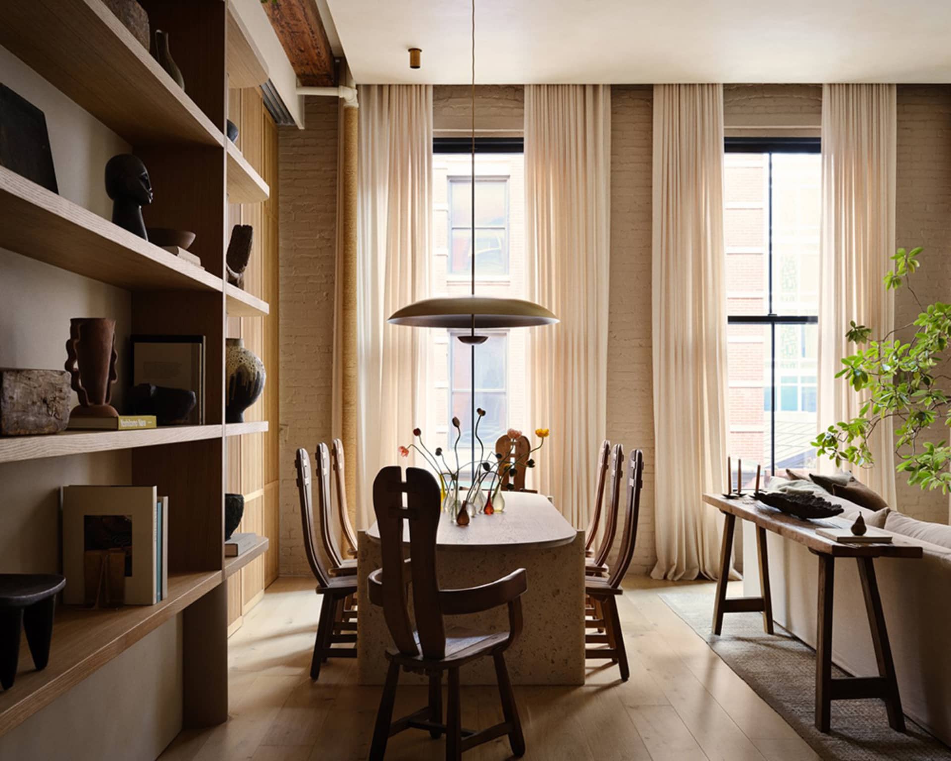 A modern dining room featuring natural wood chairs and a stone table, with a hanging pendant light and large windows with cre