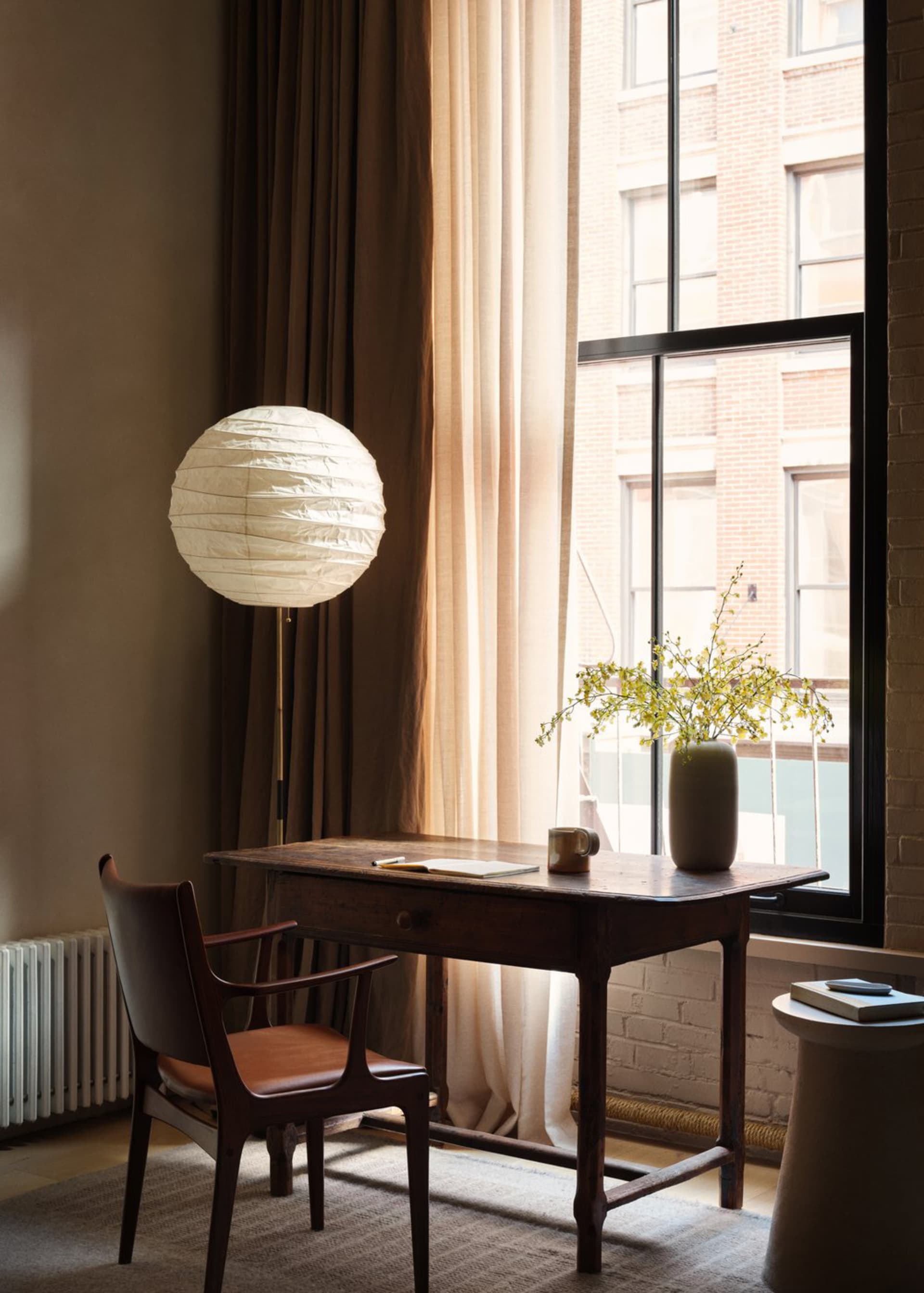 A modern home office featuring a wooden desk with a white paper lantern lamp, brown curtains, and a vase with flowers.