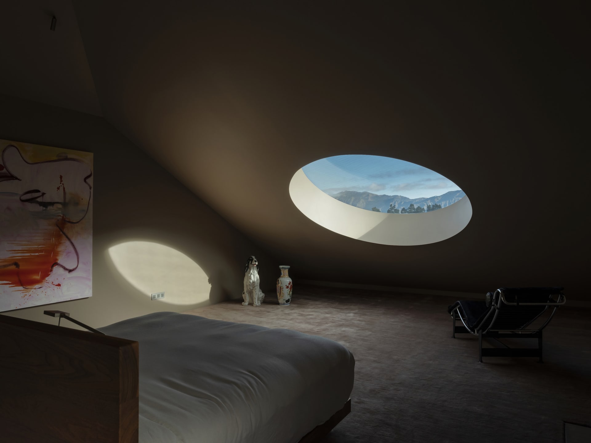 A minimalist bedroom featuring a concrete floor, white bed, and a circular window overlooking mountains.