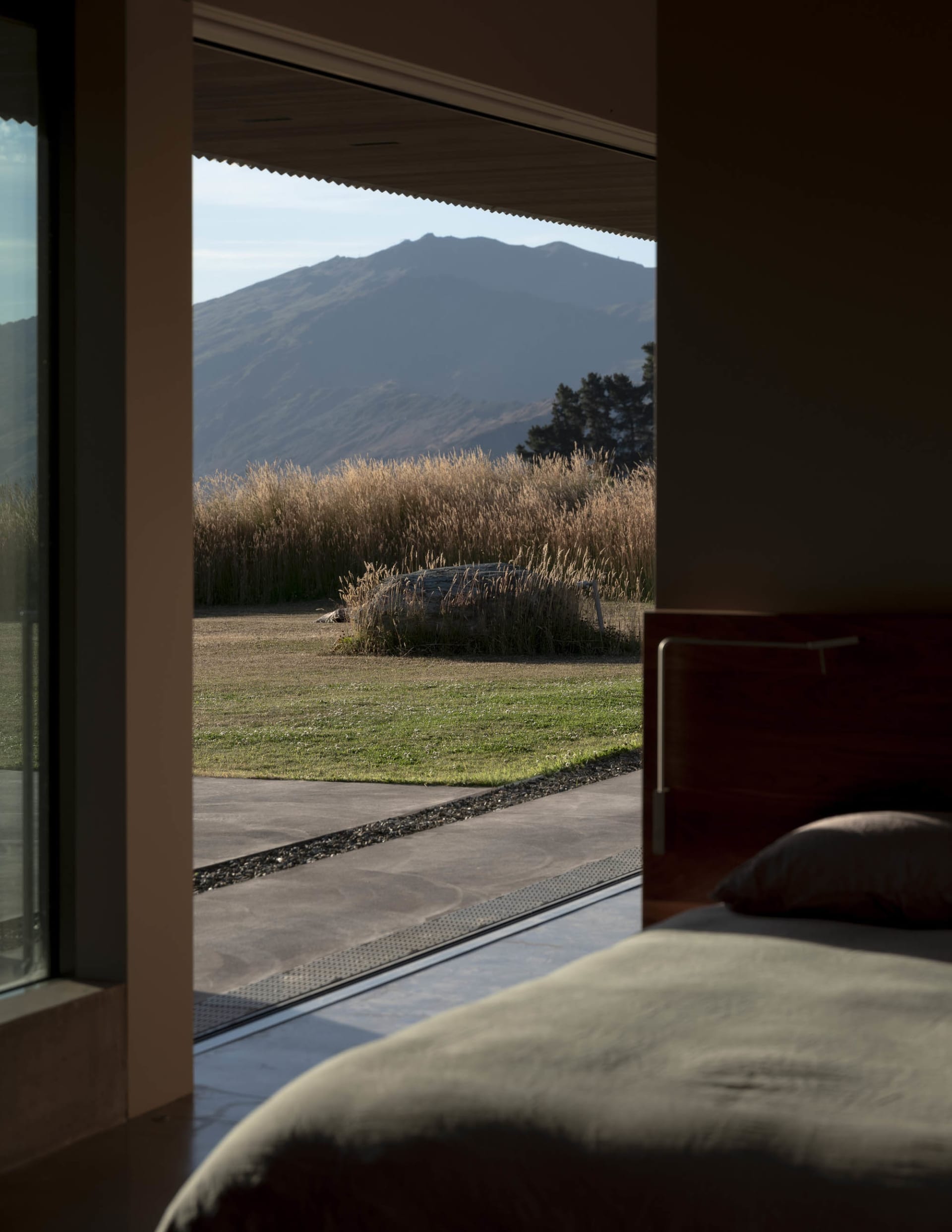 A modern residential bedroom with large windows overlooking grassy fields and distant mountains.