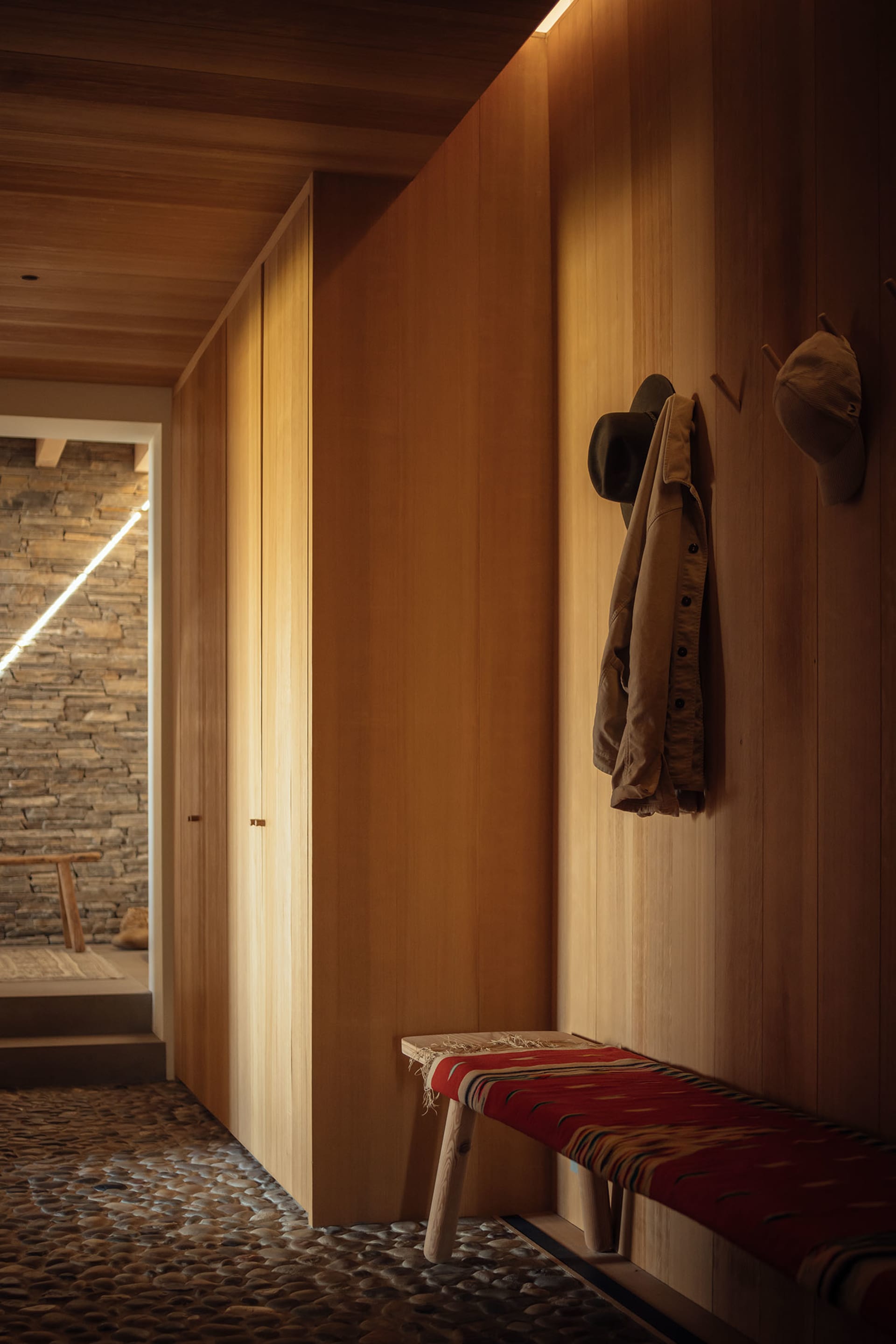 Modern entrance hall with wooden walls, wooden bench with red fabric, and stone floor.