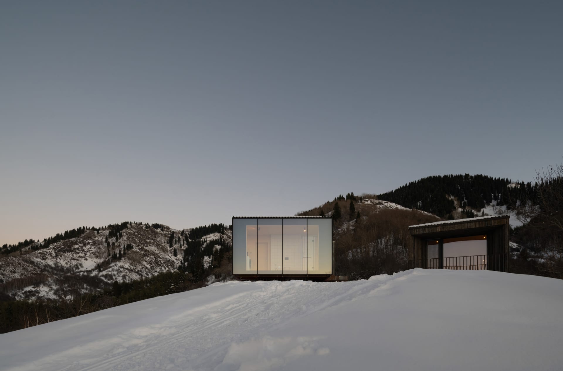 A modern, rectangular house with large glass windows on a snowy hillside.