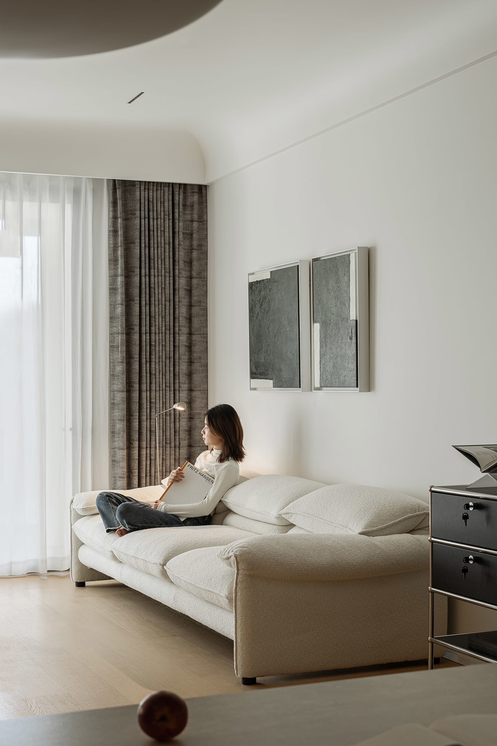 A modern minimalist living room features a white couch with plush cushions and a large window with sheer drapes, allowing nat