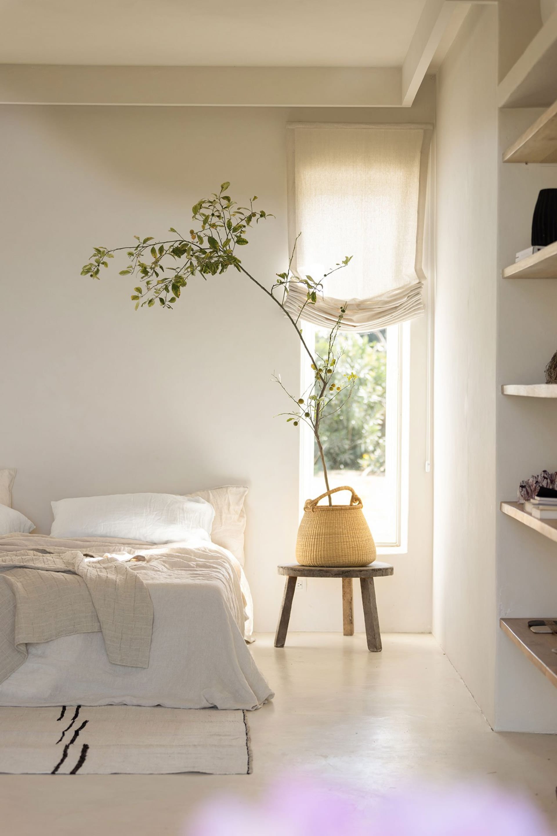 A minimalist bedroom with neutral-toned bedding, a wooden stool, and a woven basket with a plant.