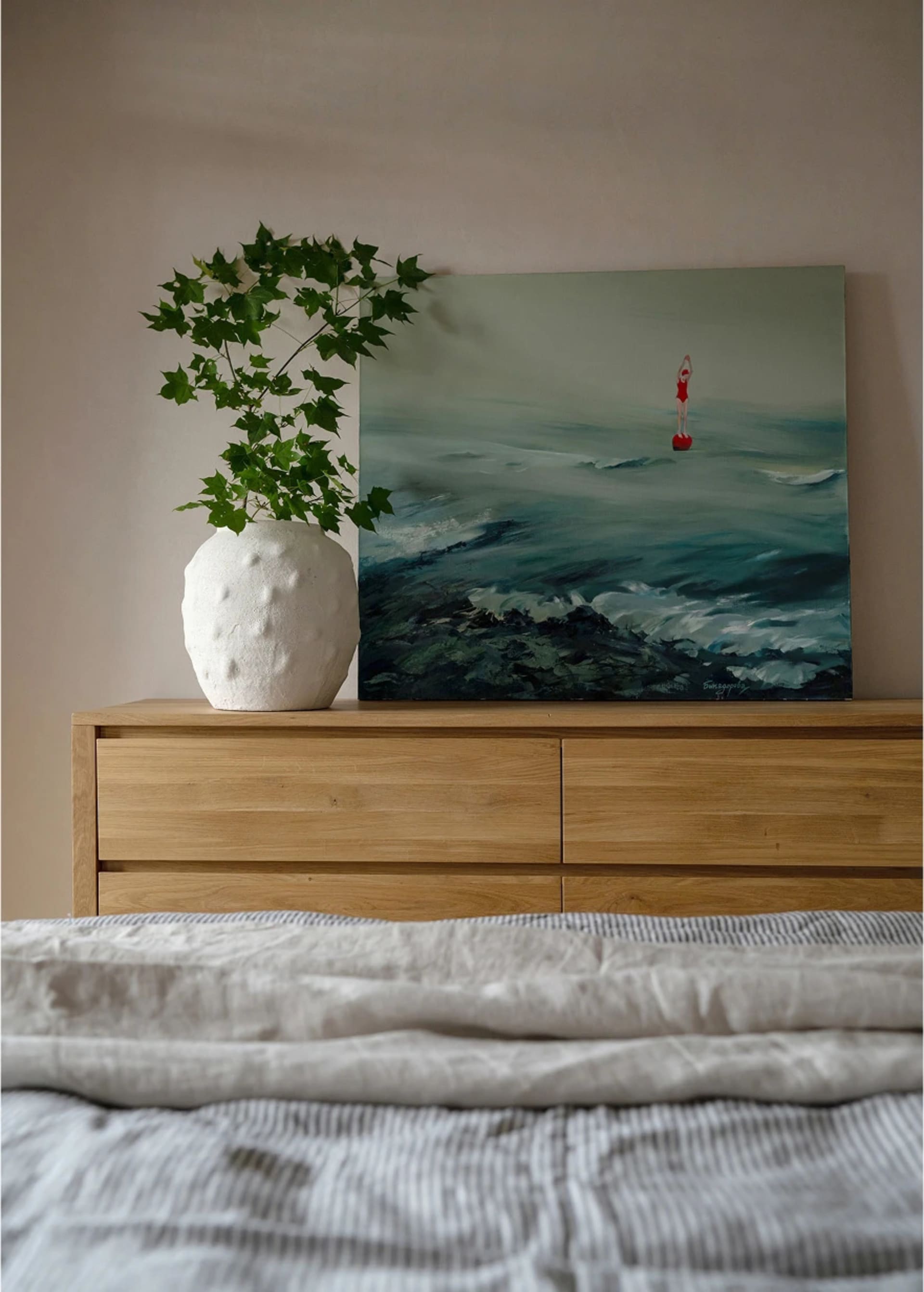 臥室內有一張木製梳妝台、白色花瓶裡的綠色植物，以及一幅描繪人在海中的畫作。