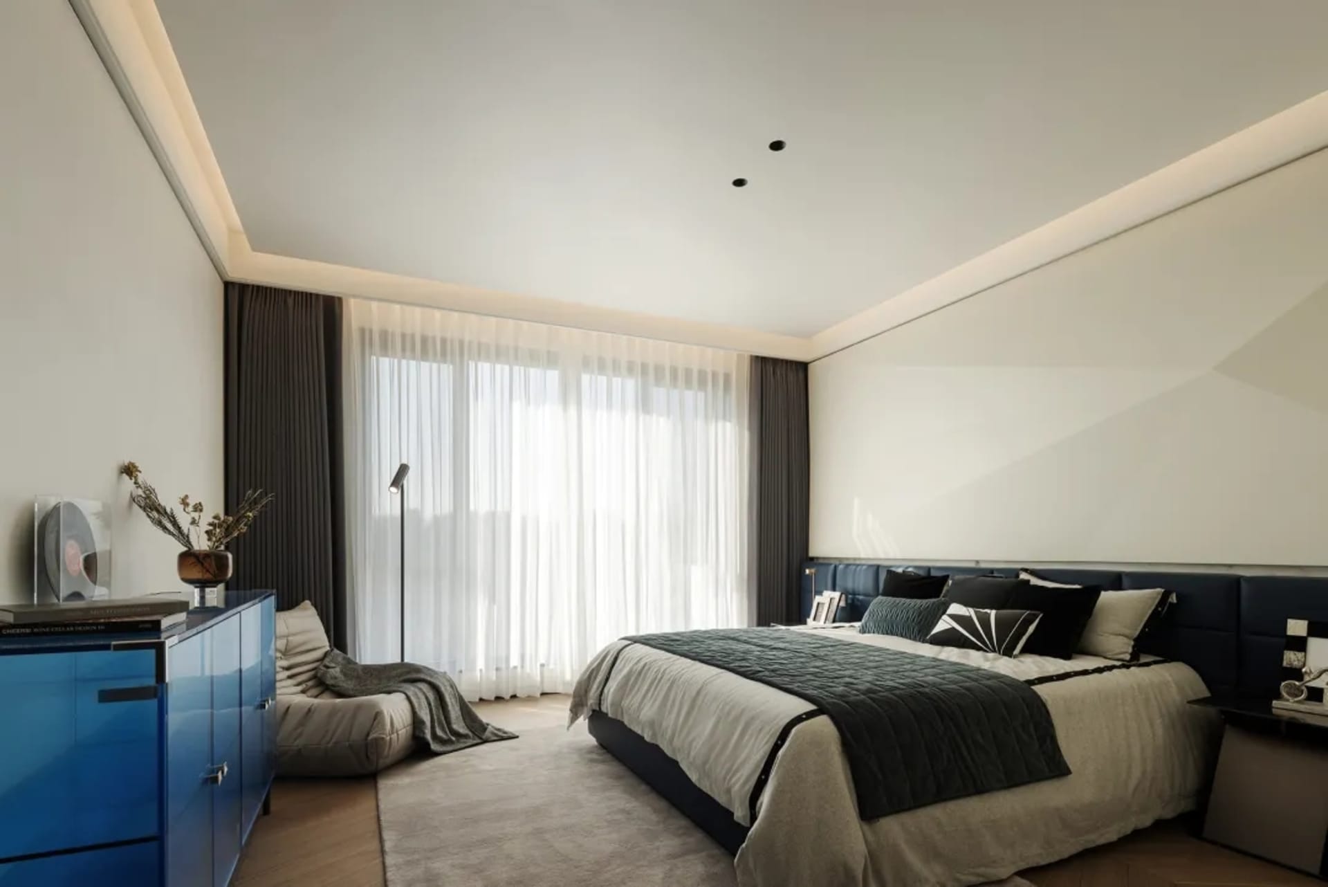 A modern bedroom featuring a large window with sheer curtains, a comfortable bed with a blue leather headboard, and a blue ca