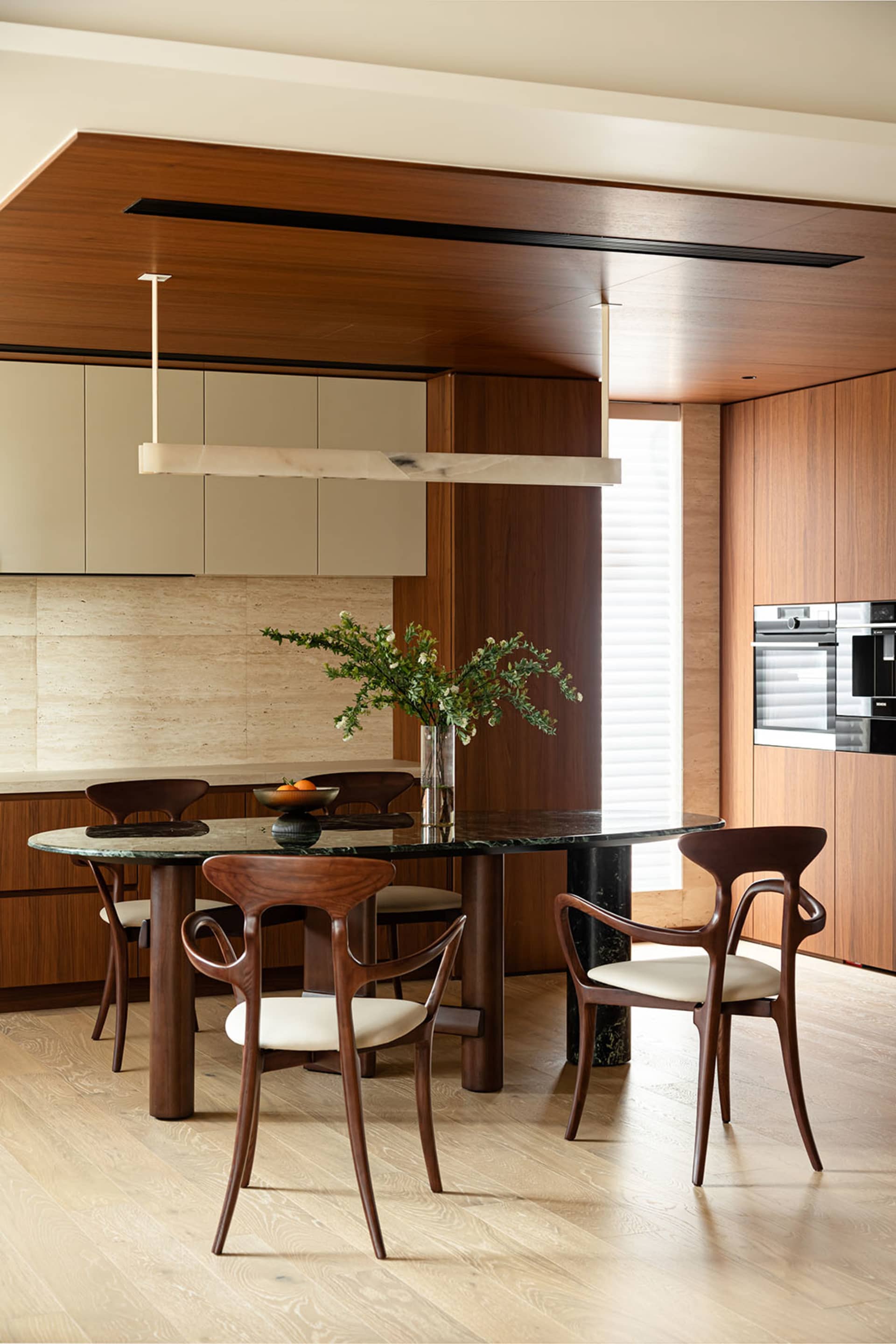 A modern kitchen with an oval dining table, wooden chairs with white upholstery, and a marble countertop. Features include wo