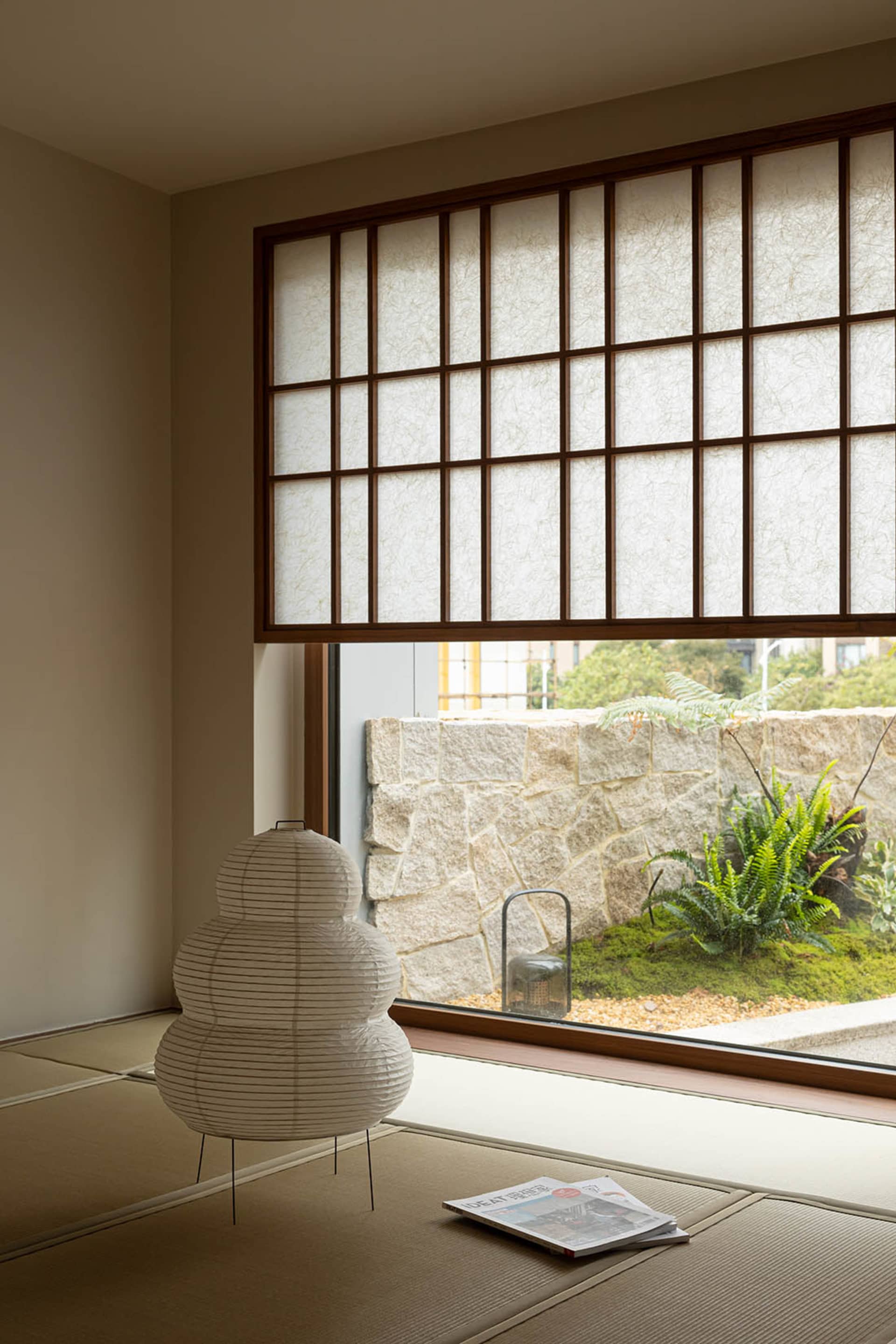 A minimalist living room with a paper-laminate floor, paper lantern, and stone wall with plants and a paper blind.