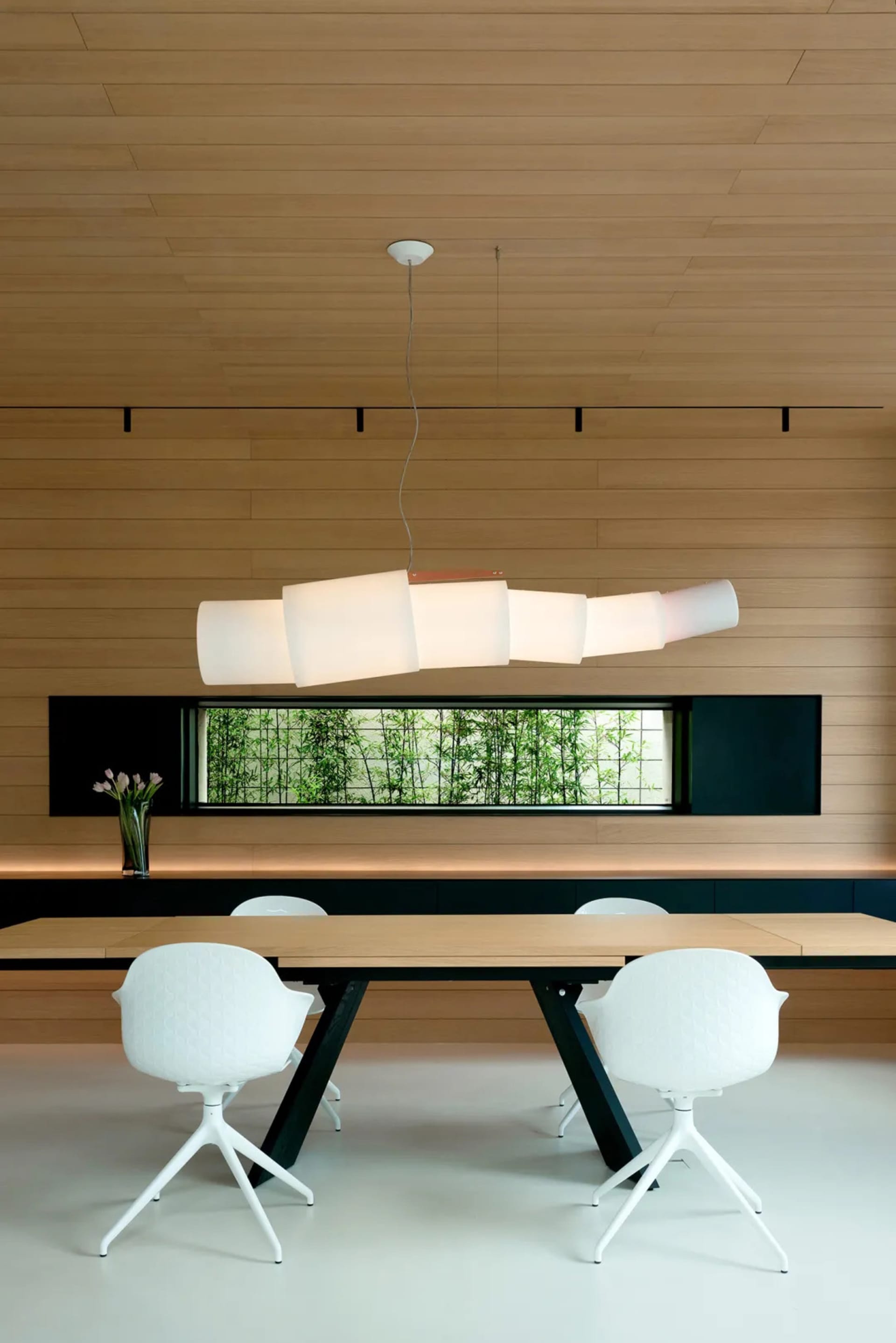 Modern dining room with white chairs and wood table, featuring a unique ceiling lamp and a window with greenery.