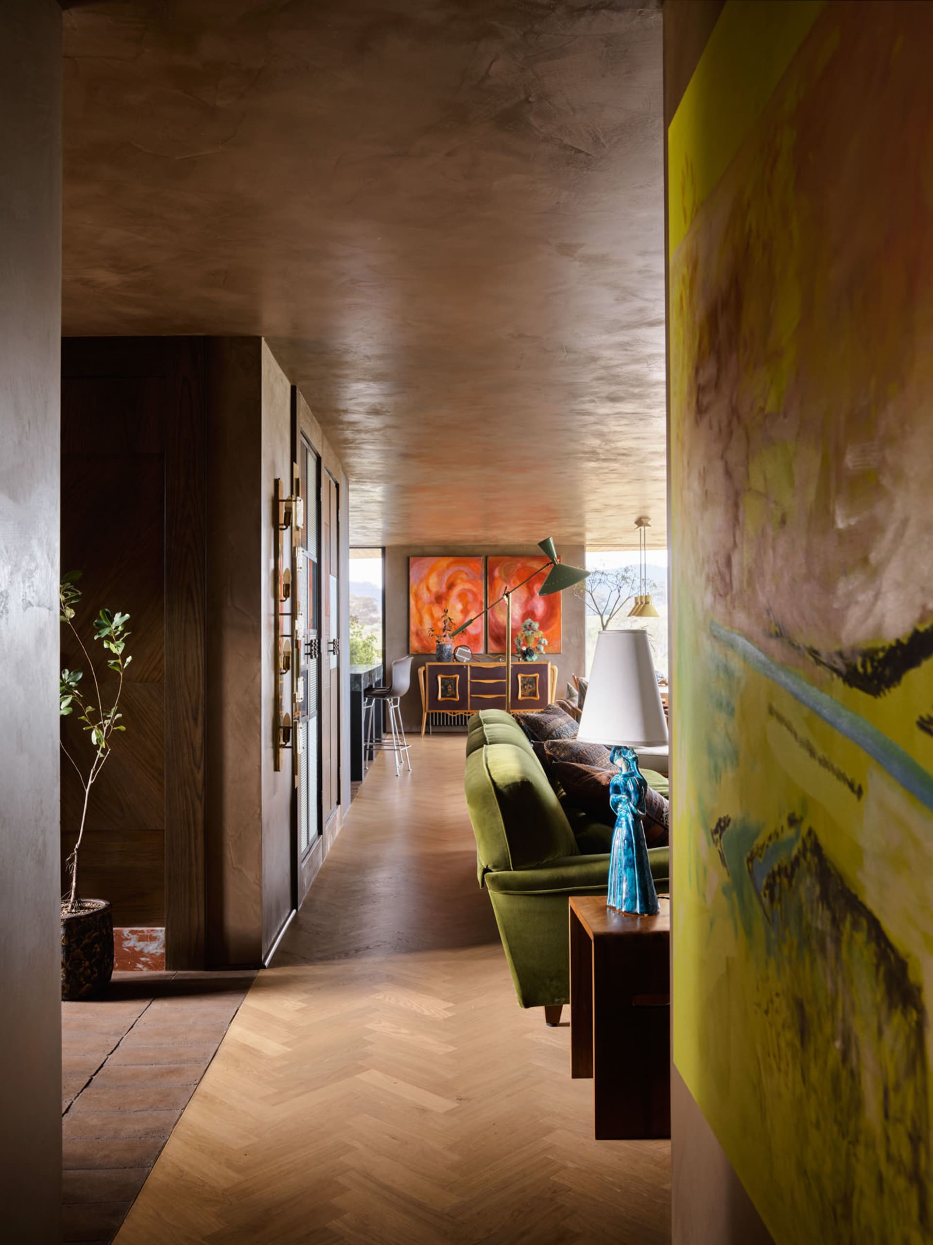 A modern residential living room with concrete ceiling and wooden flooring, featuring a green sofa and abstract paintings.