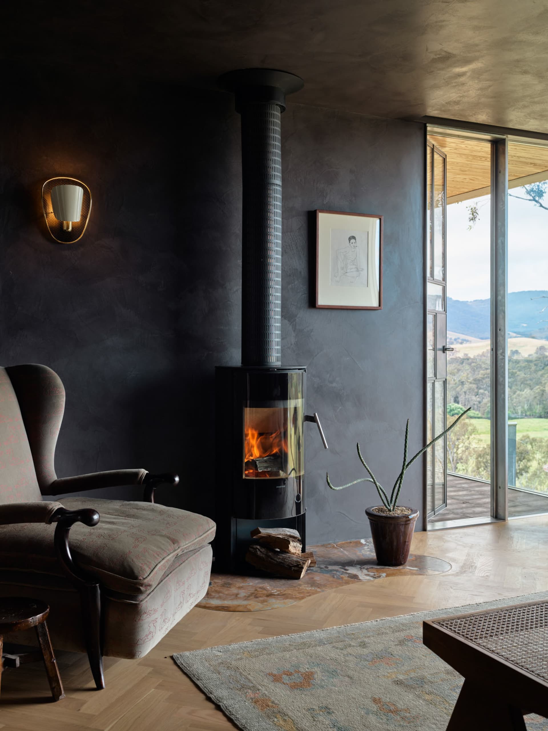 A cozy industrial-style living room features a concrete fireplace, wood floors, and a rustic rug, with a view overlooking a g