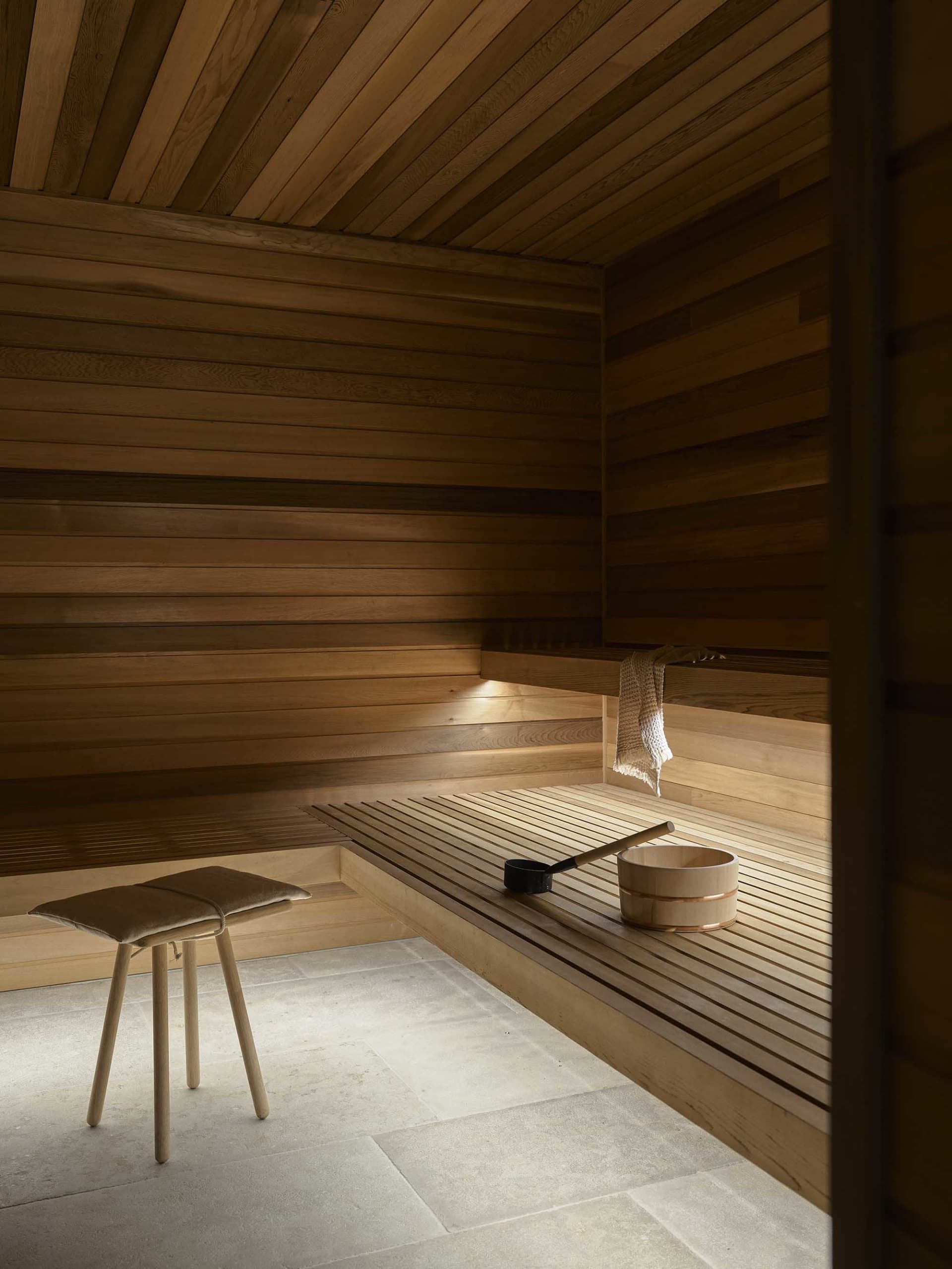 An industrial sauna room with wooden walls and benches, featuring a stool and a wooden bucket with a ladle.