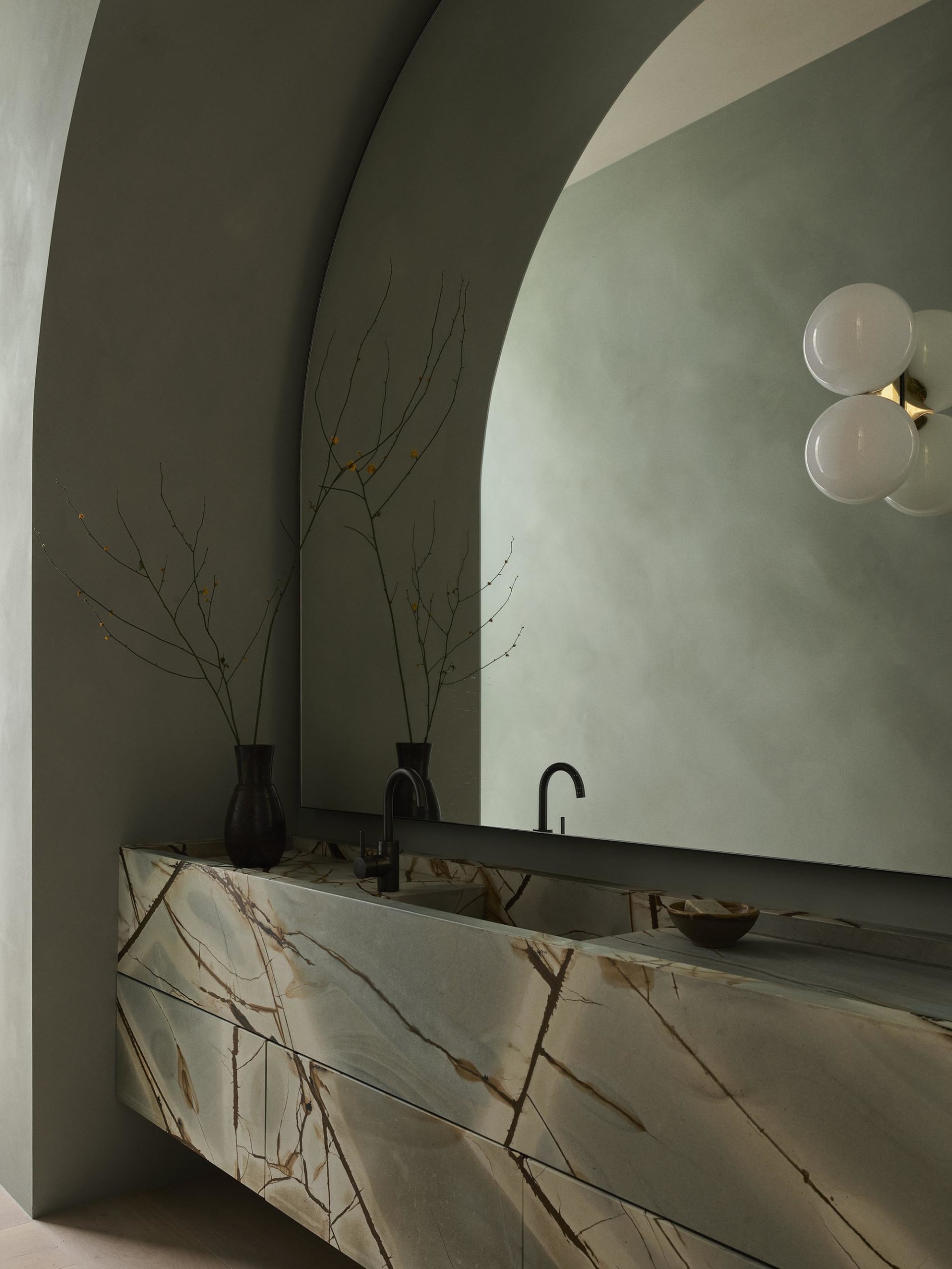 A sleek, modern bathroom featuring a marble vanity, cylindrical light fixture, and a large, arched mirror.