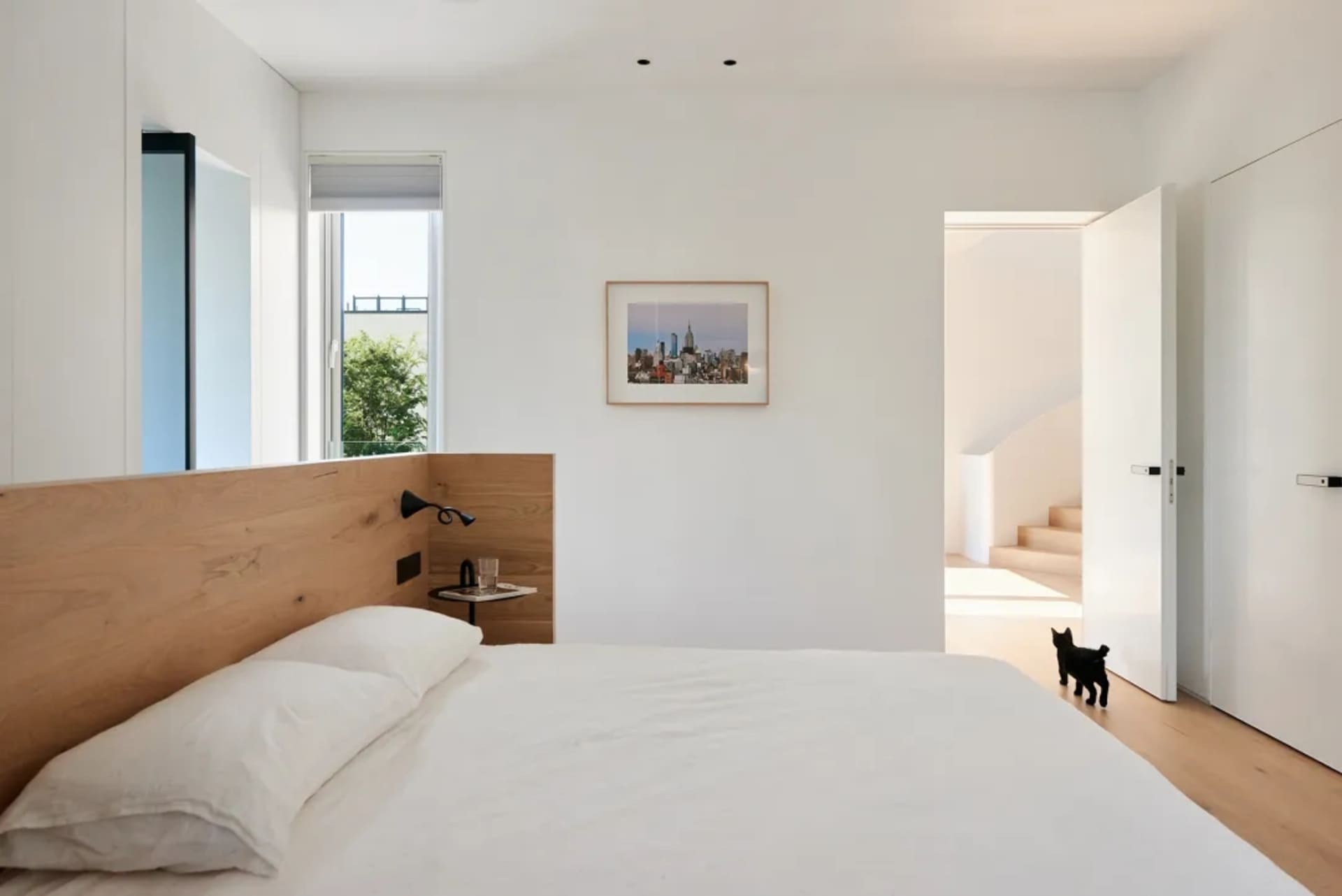 Modern minimalist bedroom with wooden headboard, white bedding, cityscape artwork, and black cat.