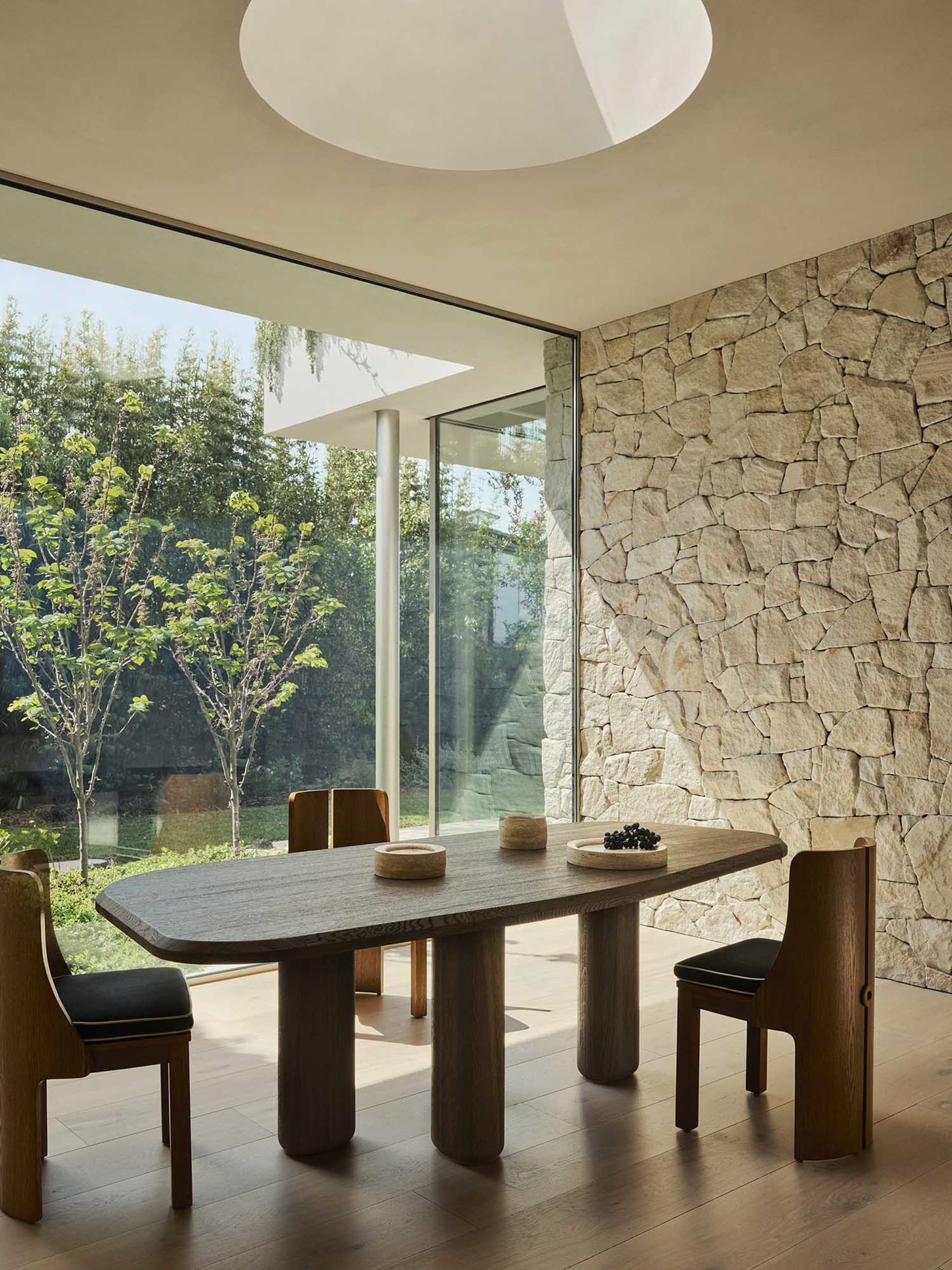 A minimalist dining room with a wooden table and a stone wall, featuring floor-to-ceiling windows that allow natural light to
