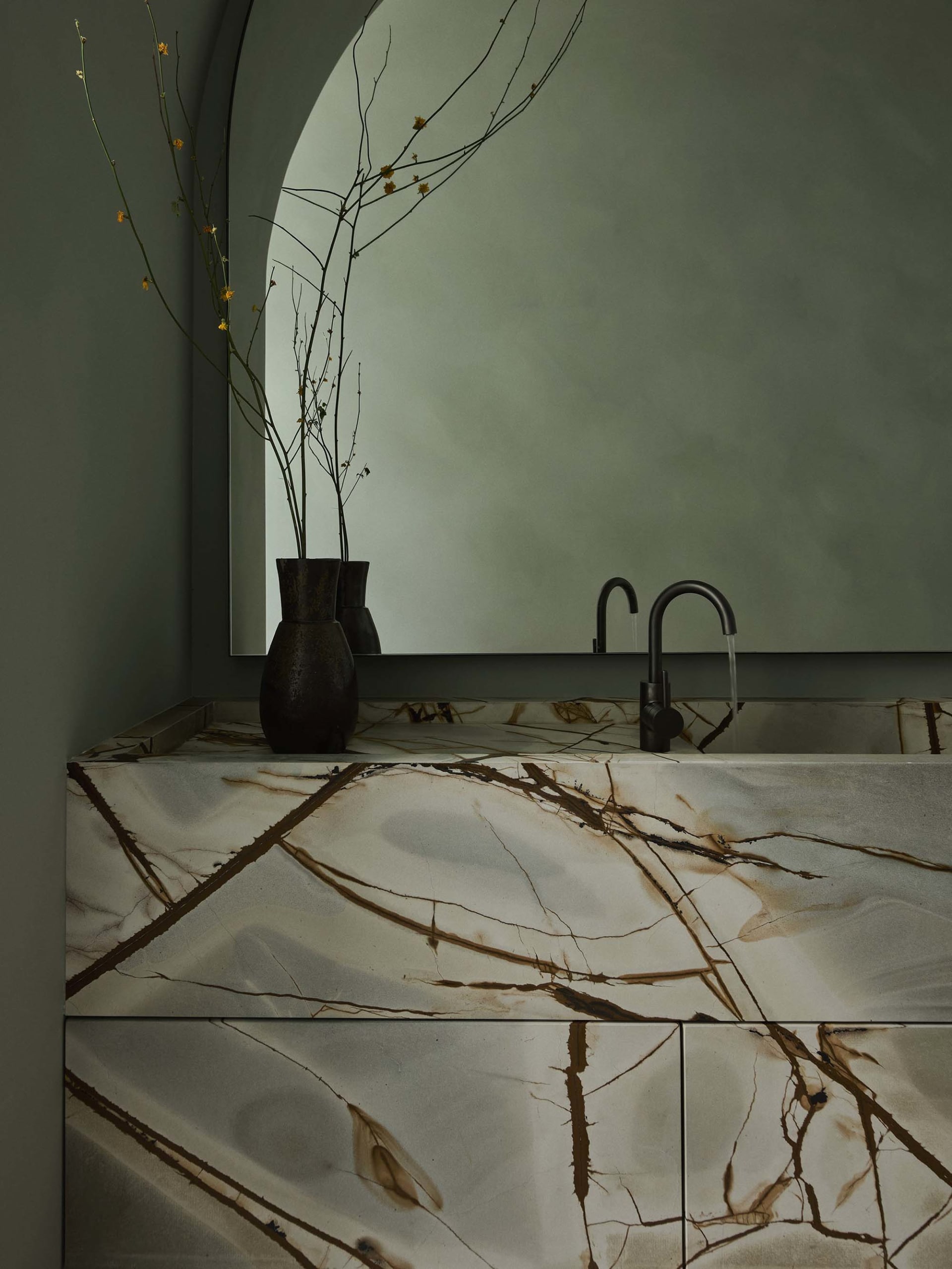 Modern bathroom with a marble countertop featuring dark veins, a black faucet, and a vase with dried branches on a circular w