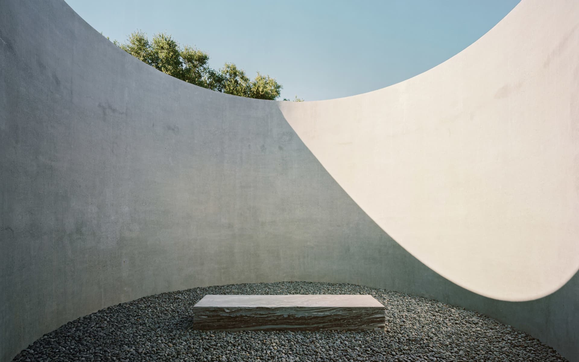 A minimalist concrete sculpture garden featuring a smooth, rectangular stone pedestal surrounded by pebbles and set against c