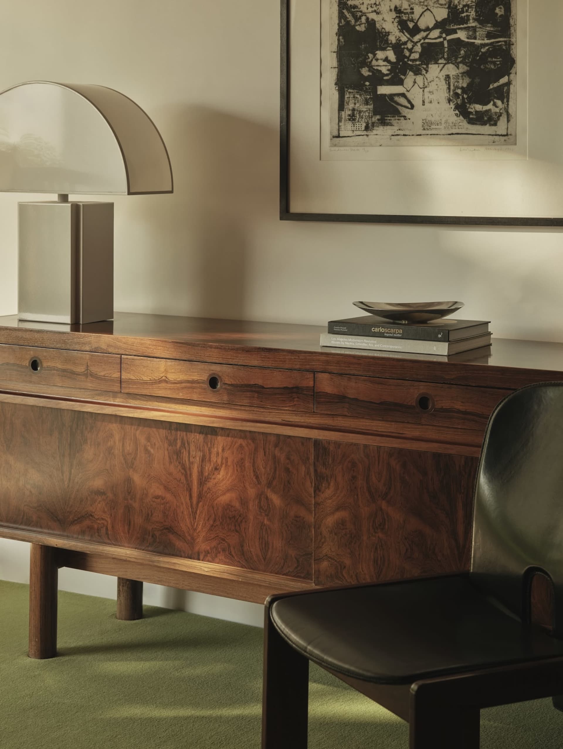 A modern office desk made of wood with a lamp, chair, and framed artwork.