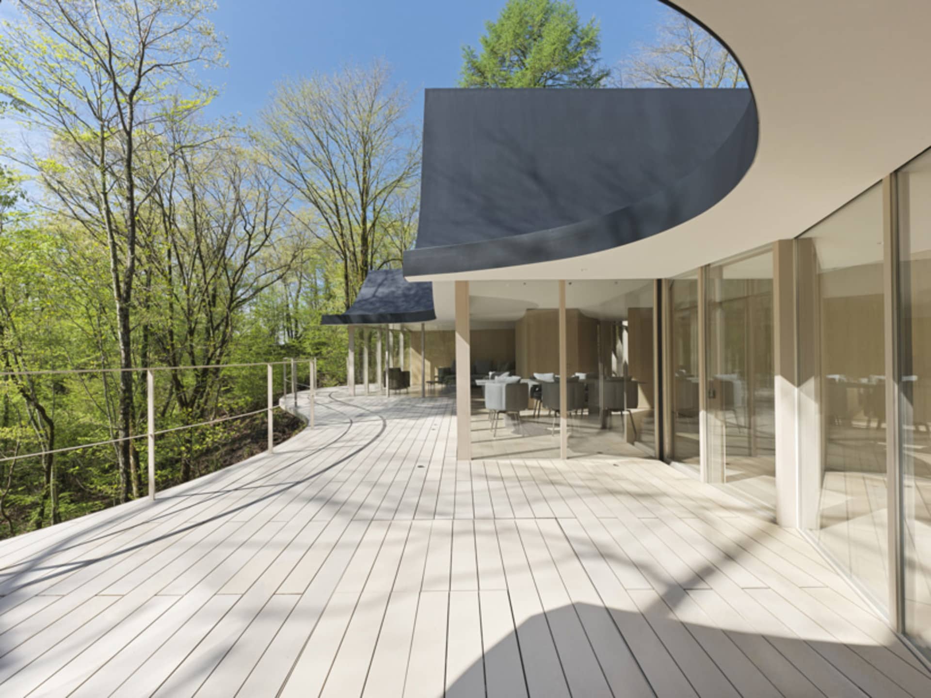 Modern residential deck featuring a dining table and chairs with wood flooring and glass railings surrounded by greenery.