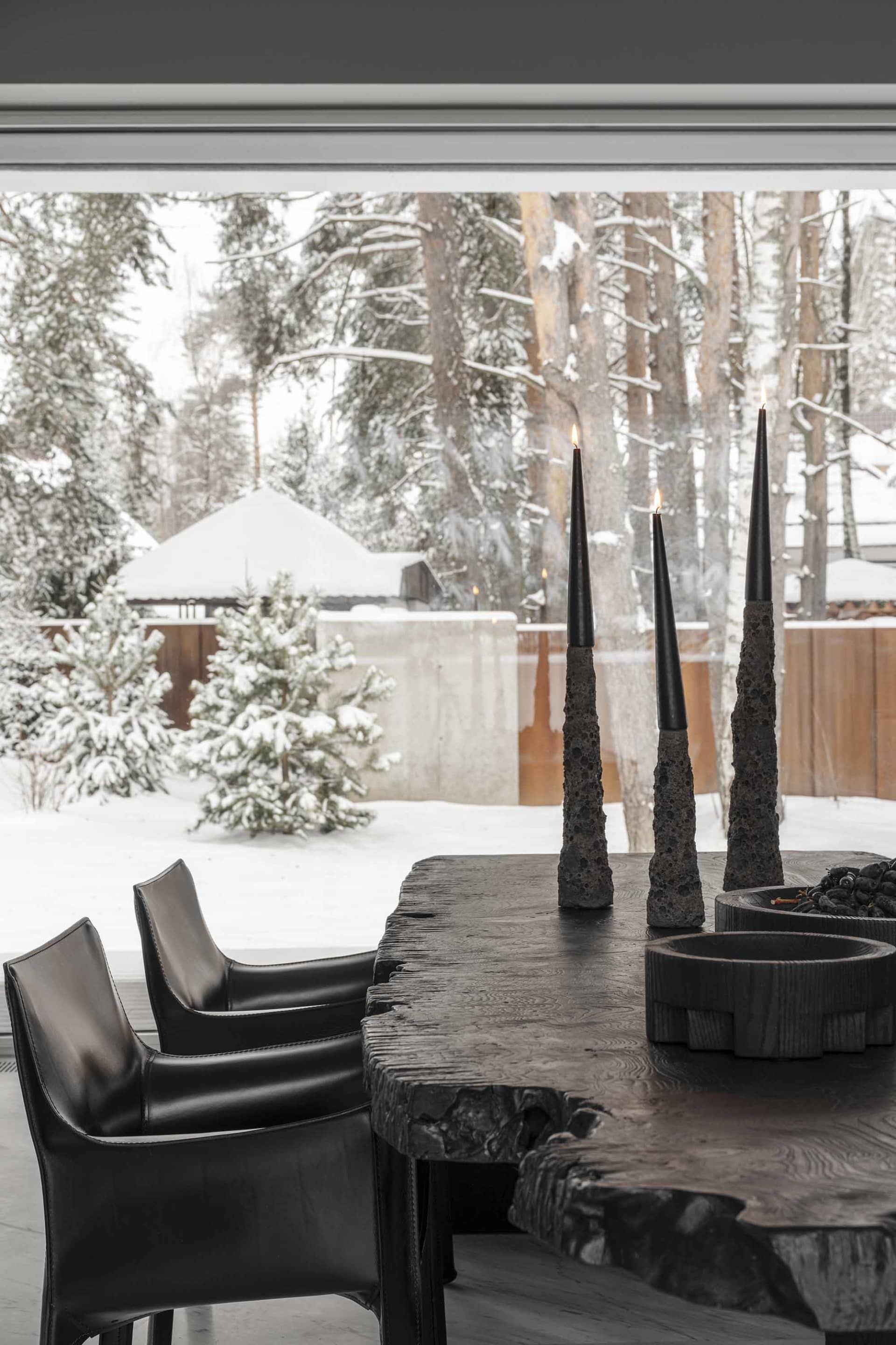 A cozy winter cabin scene with snow-covered trees, a black leather chair, and black candles on a wooden table.
