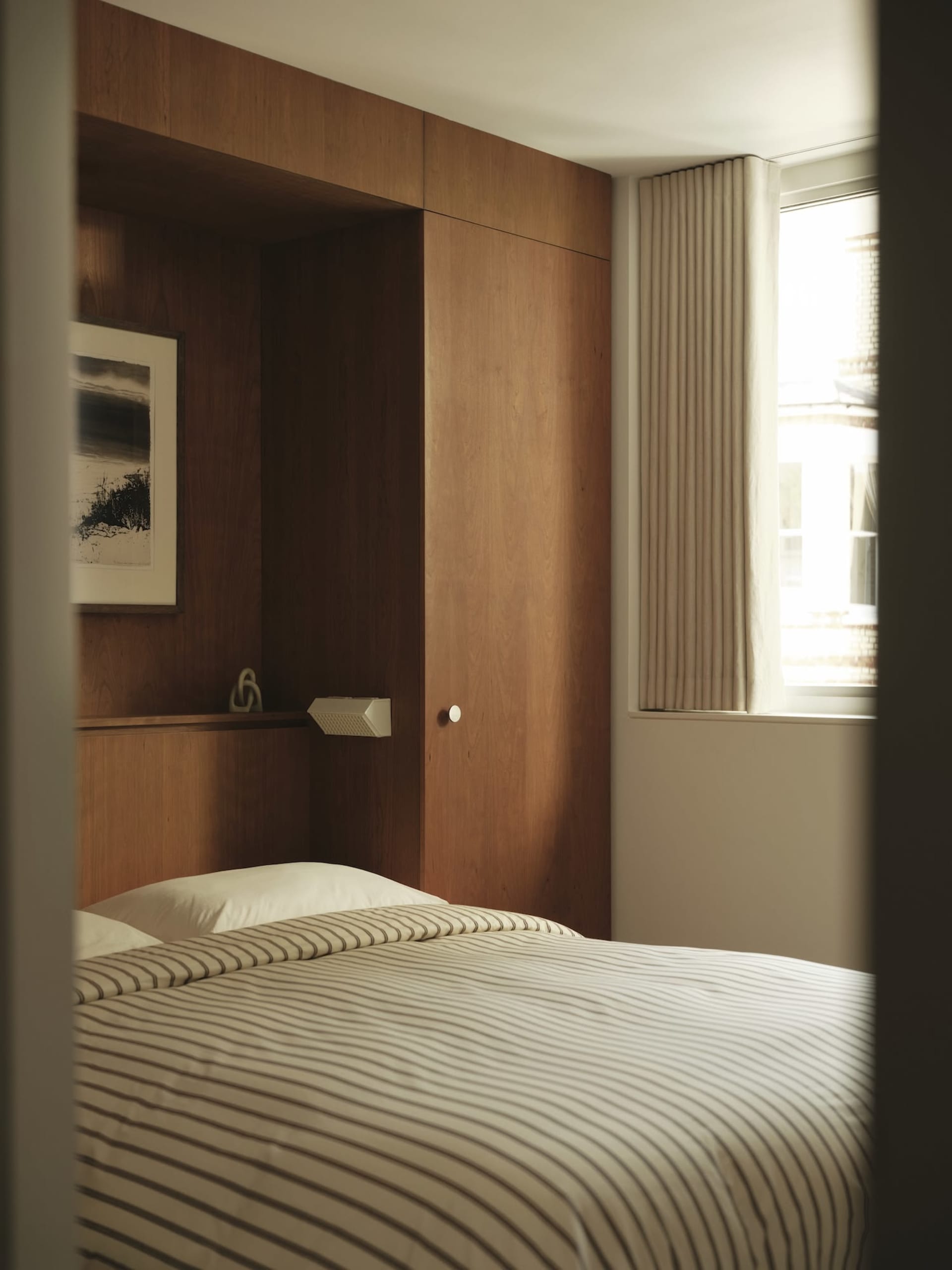A modern minimalist bedroom featuring a wooden paneled wall, white bedding with gray stripes, and a window with beige curtain
