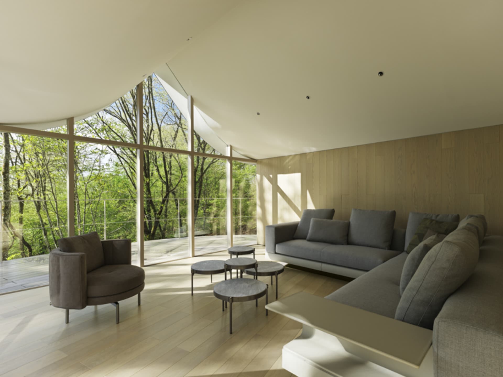 A modern minimalist living room featuring a large sofa, coffee tables, and floor-to-ceiling windows with a view of green tree
