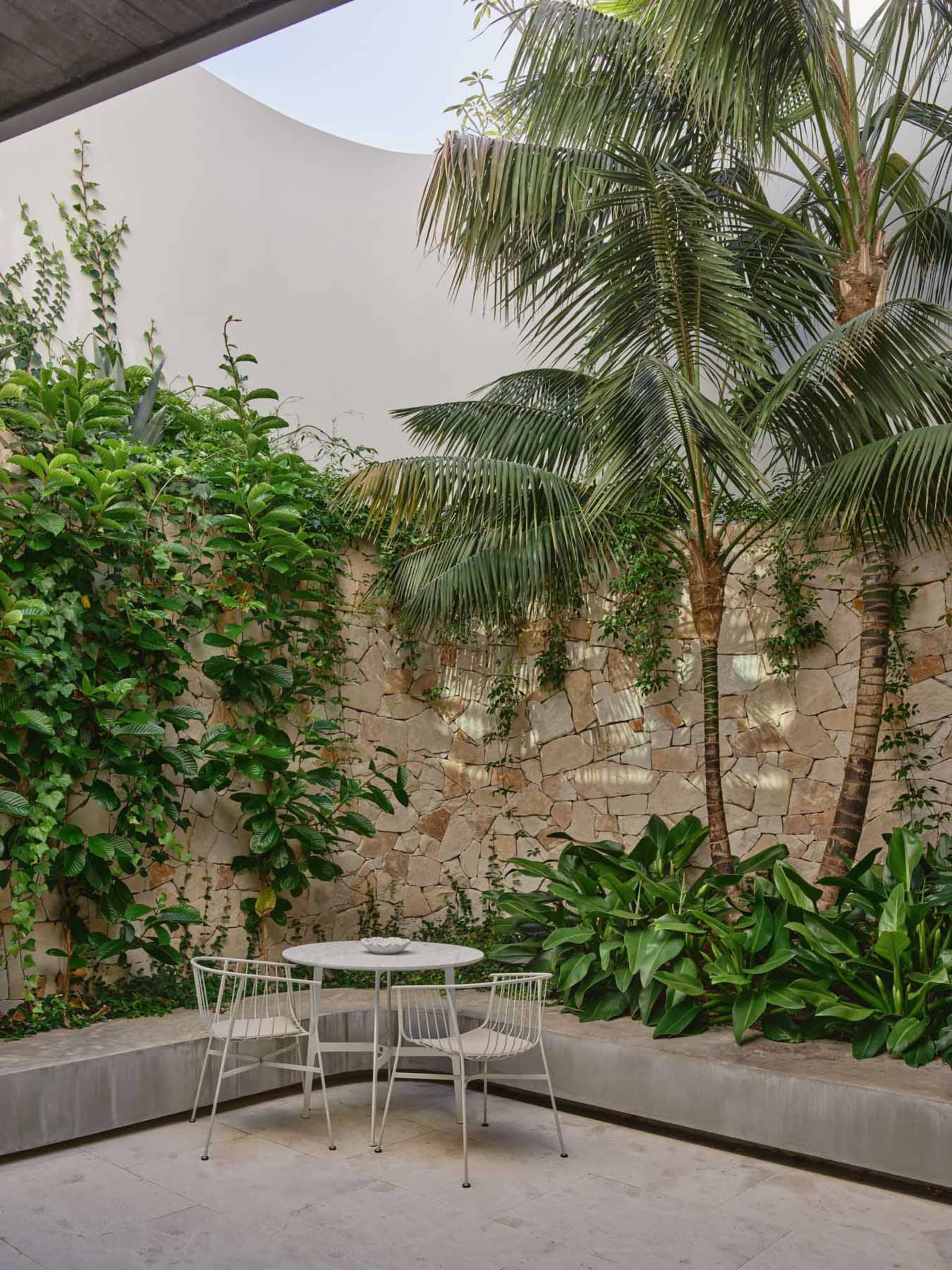 A modern outdoor space featuring a circular white table and chairs on a concrete patio surrounded by greenery and palm trees.