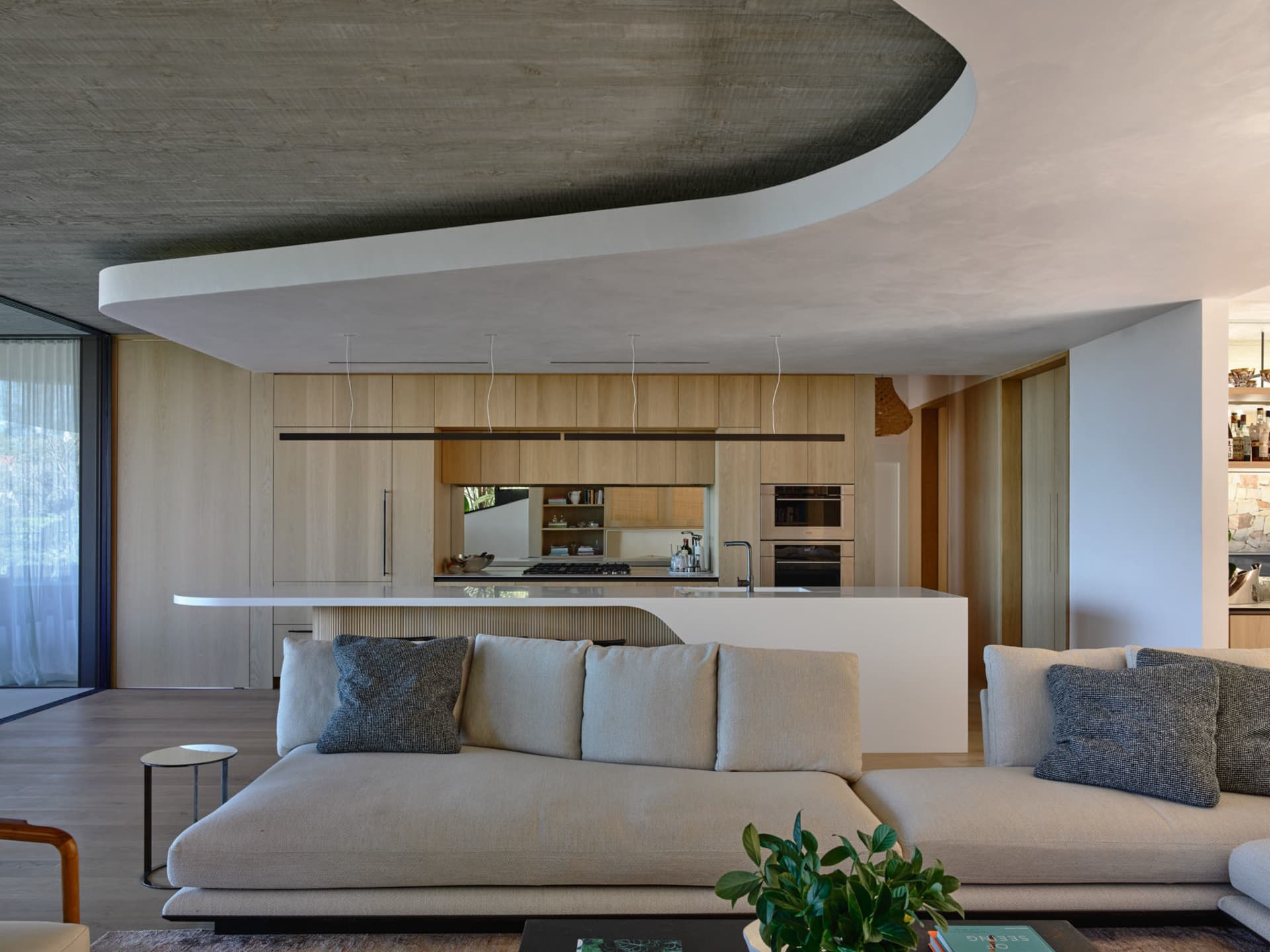 Modern living room with a concrete ceiling and curved design, featuring a beige sectional sofa with grey pillows, a green pla