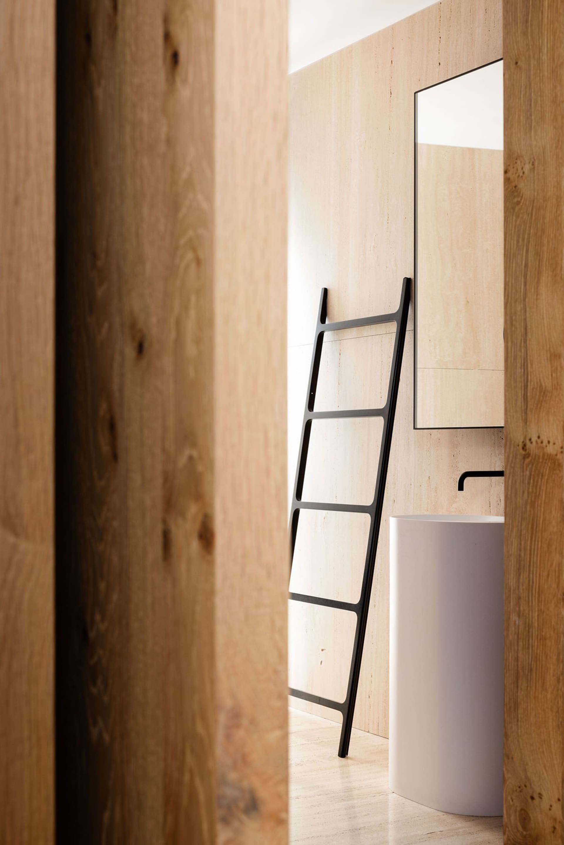 Modern minimalist bathroom featuring a wooden ladder, marble sink, and metal fixtures against a beige marble wall.