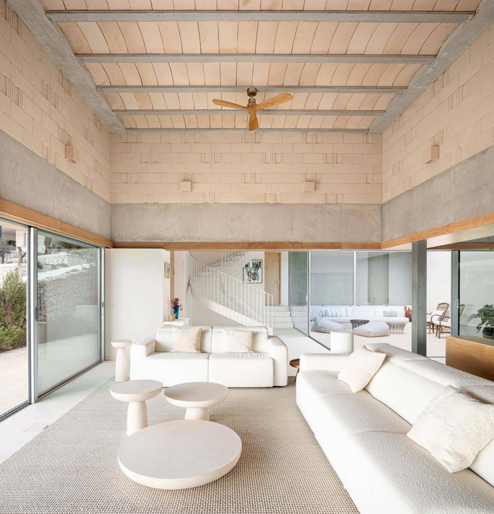 A modern living room with concrete walls and ceiling, featuring white sofas, white coffee tables, and a spiral staircase.