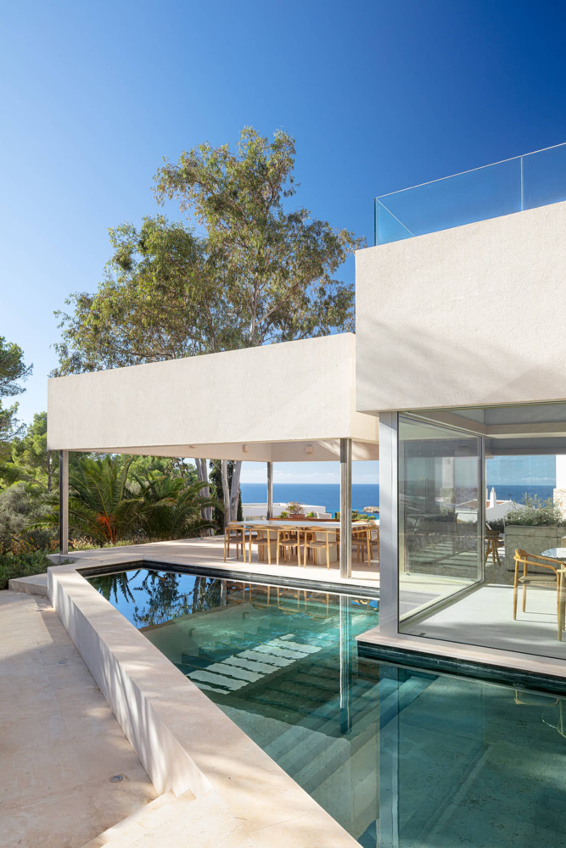 Modern residential exterior featuring a glass wall and concrete patio with a swimming pool and dining area.