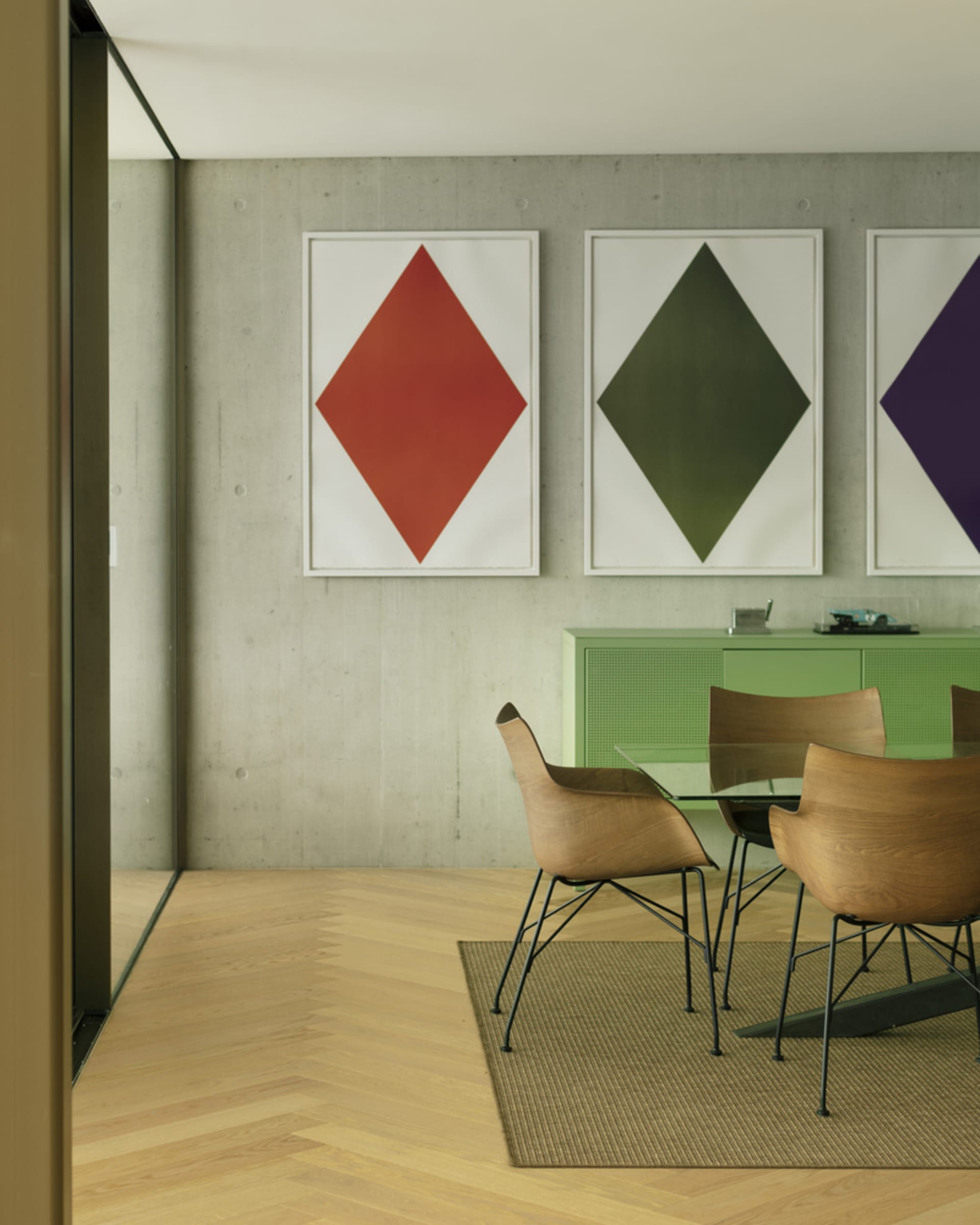 A modern dining room featuring wooden chairs, a glass table, metal legs, and geometric artwork on a concrete wall.