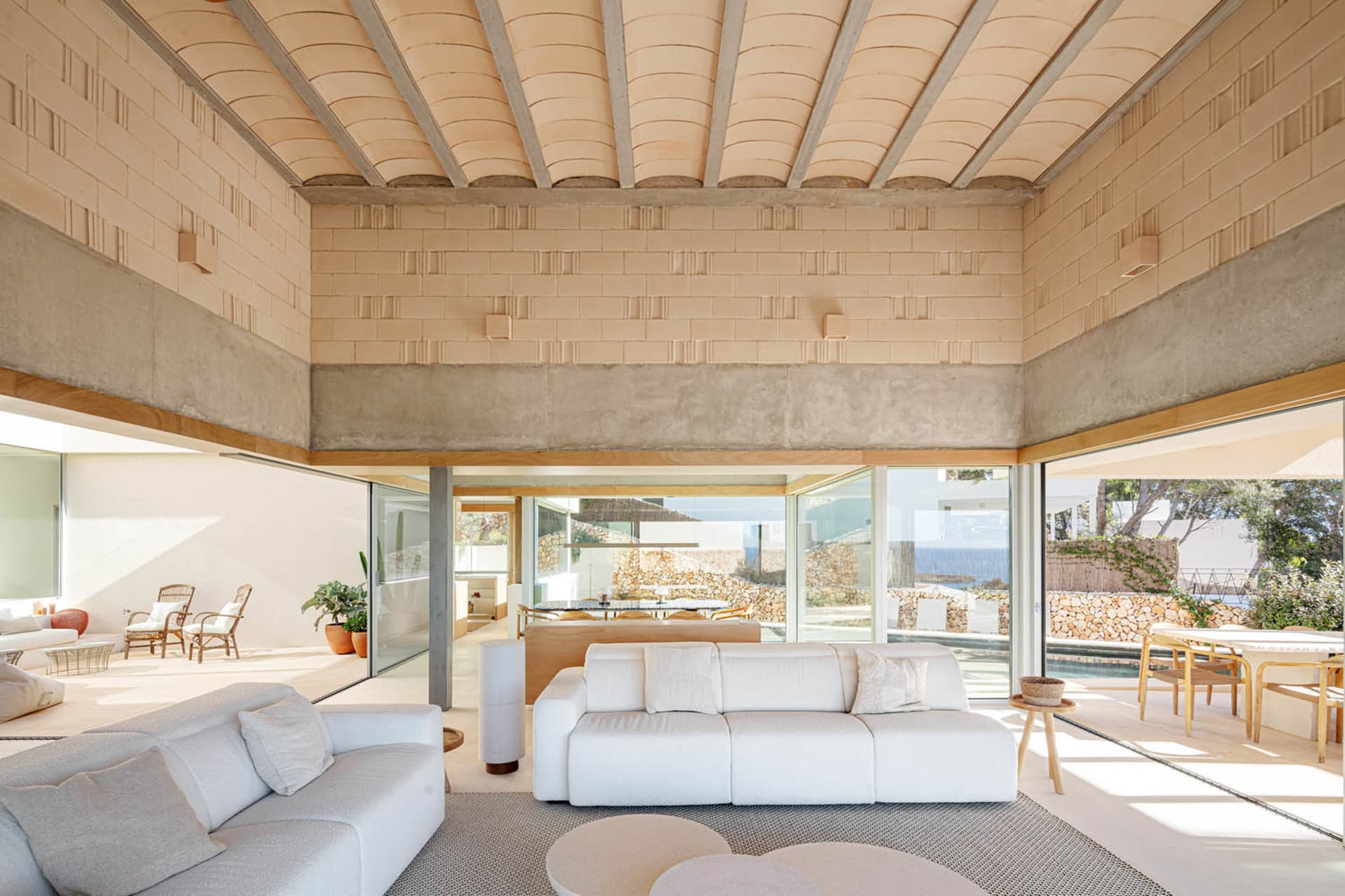 A modern living room with a concrete floor and walls, featuring white sofas and a high ceiling with wooden beams.