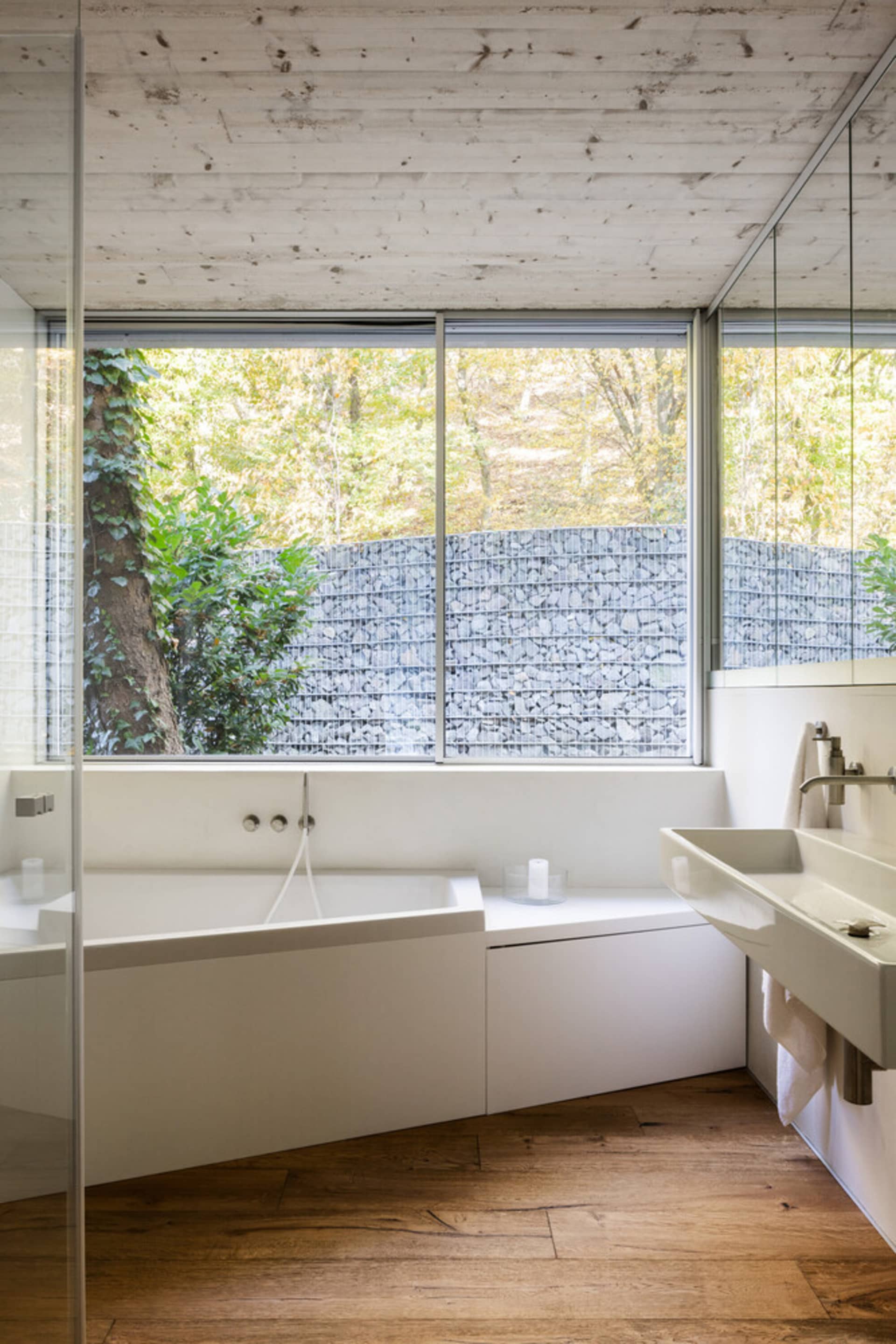A minimalist bathroom interior with a white stone bathtub and sink, large window overlooking a forested area, and wooden floo