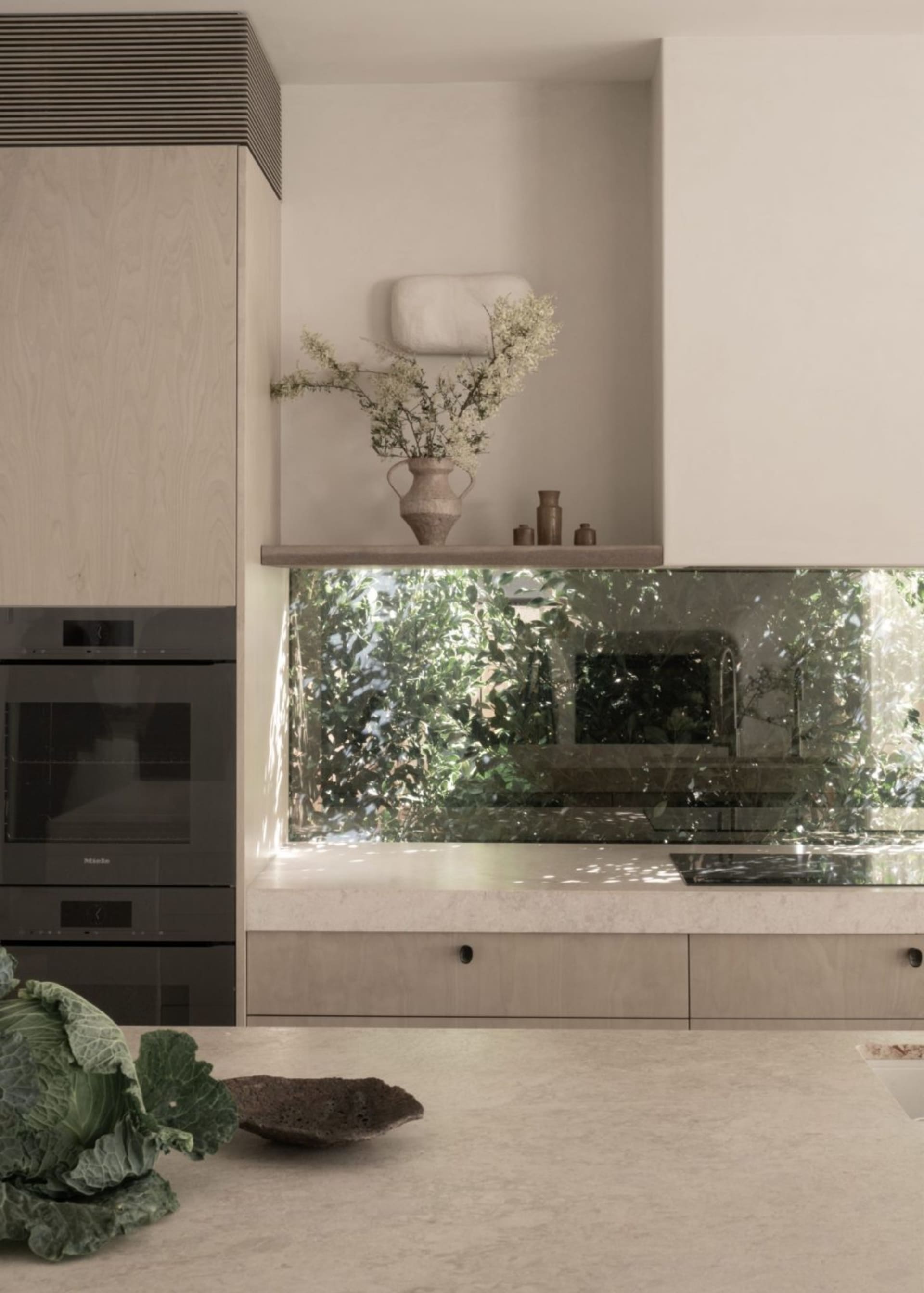 A modern, minimalist kitchen with taupe cabinets and a reflective backsplash showcasing greenery.