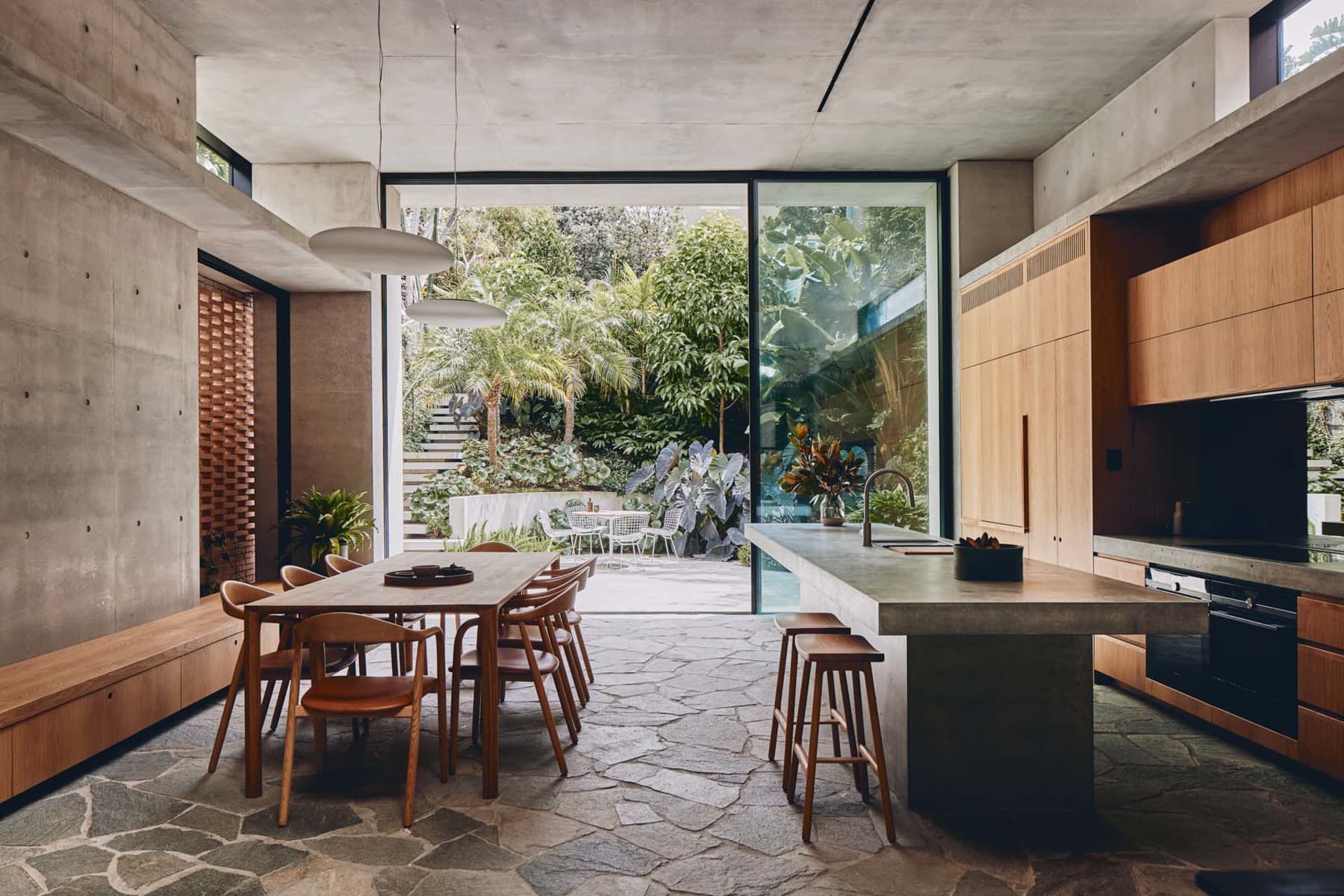 A modern kitchen featuring concrete and glass materials with a stone floor and large windows looking out to a garden.