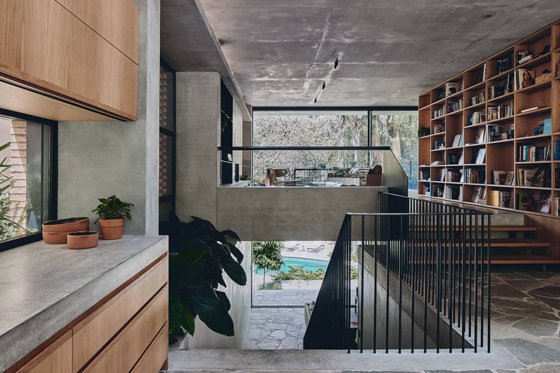 A modern residential library with concrete walls and wooden shelves, featuring a stairway leading to another level.