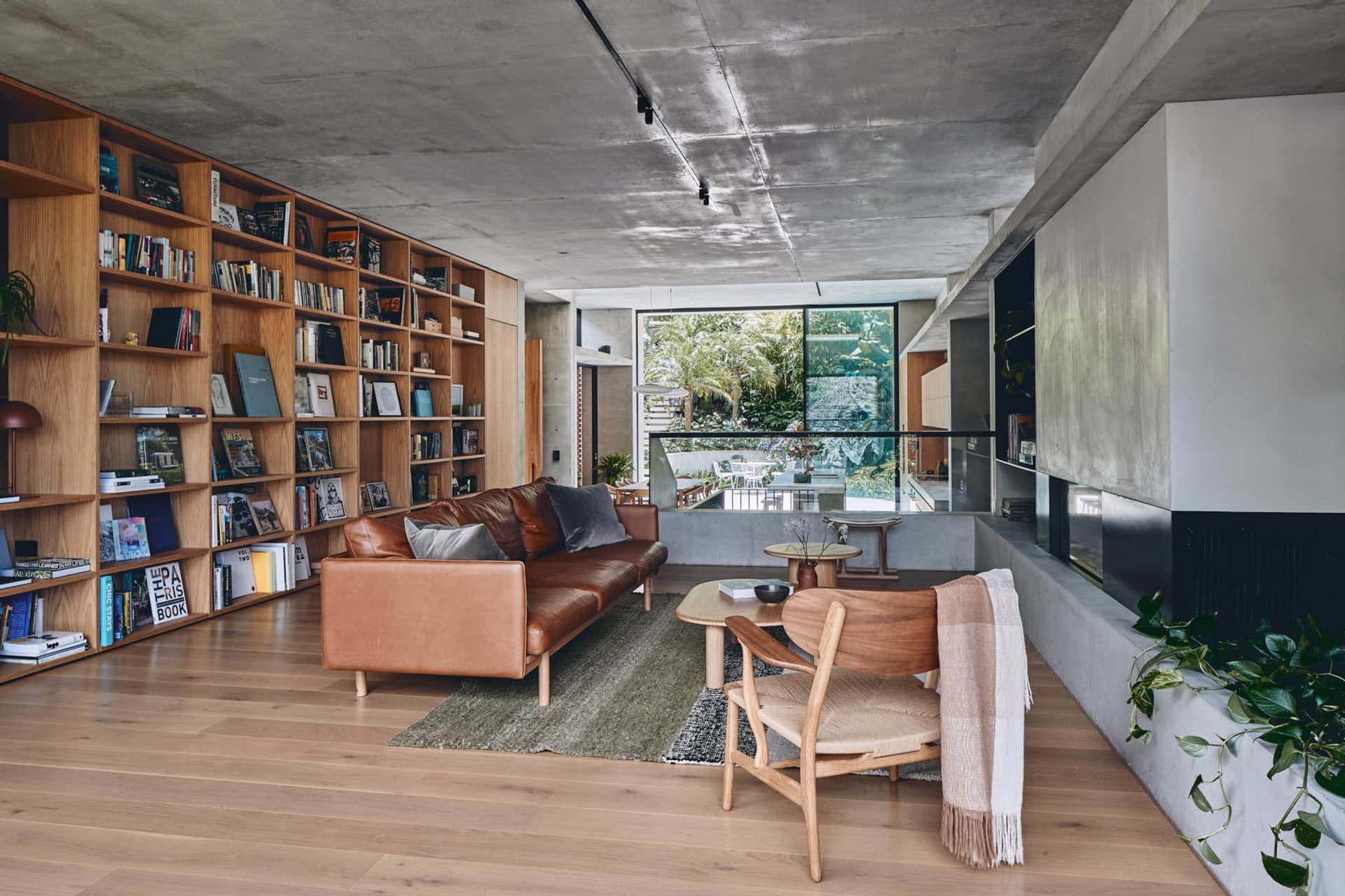A modern living room with concrete walls, wooden furniture, and a large bookshelf filled with books and various items.