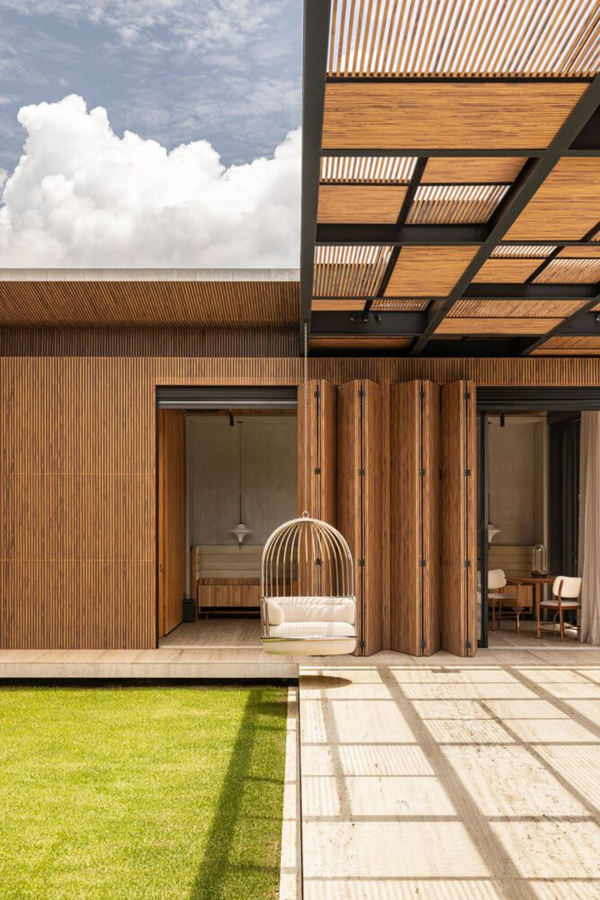 Modern residential exterior featuring wooden elements, concrete surfaces, and glass windows under a clear blue sky with fluff