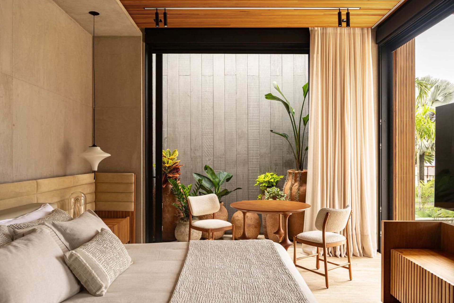 A minimalist bedroom with a bed, chairs, and a small table surrounded by potted plants, featuring hardwood floors and fabric