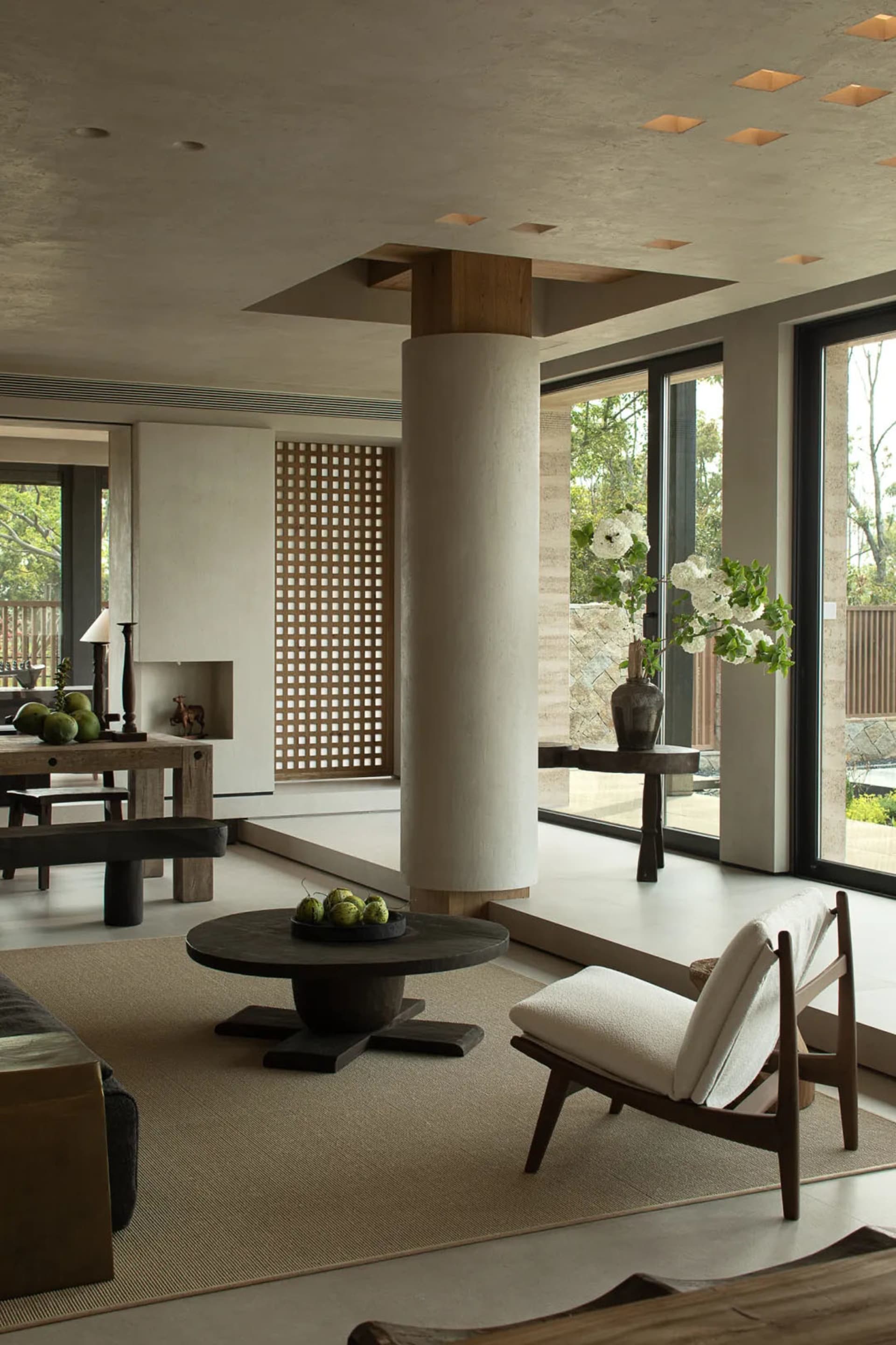 A modern minimalist living room featuring concrete and wooden elements with a round table and floor-to-ceiling windows.