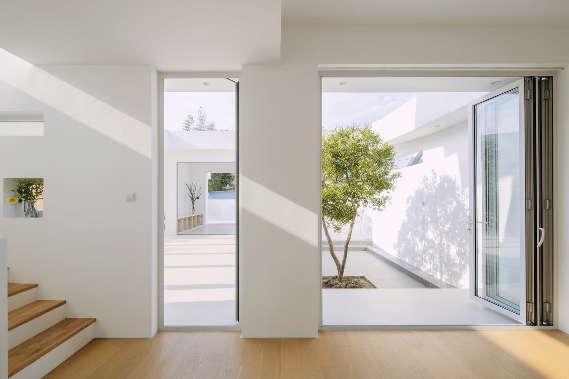A minimalist white interior space with a large glass window and wooden floor, featuring a tree in the background.