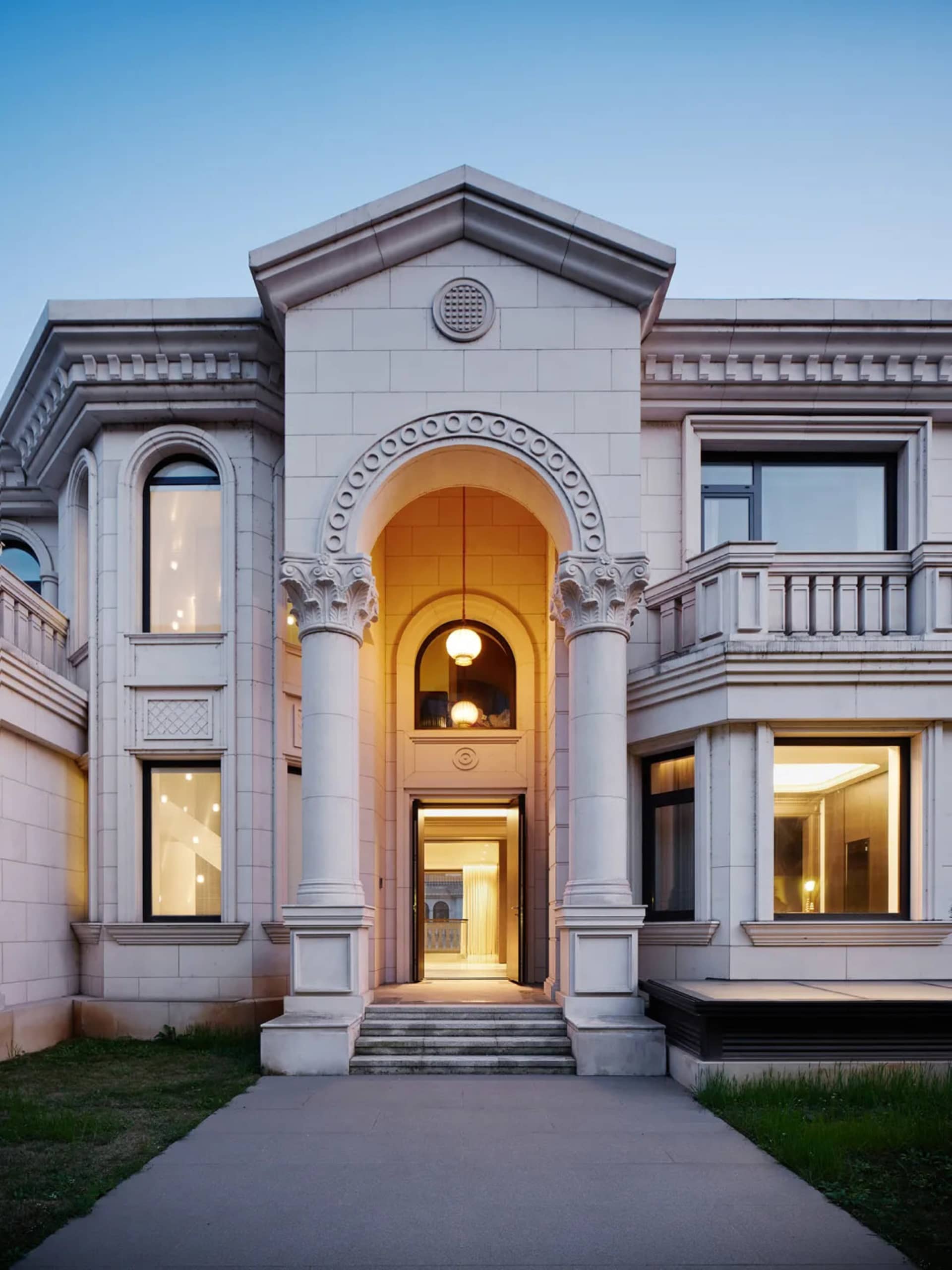 A modern house with a stone archway entrance during dusk with interior lights on and clear sky.