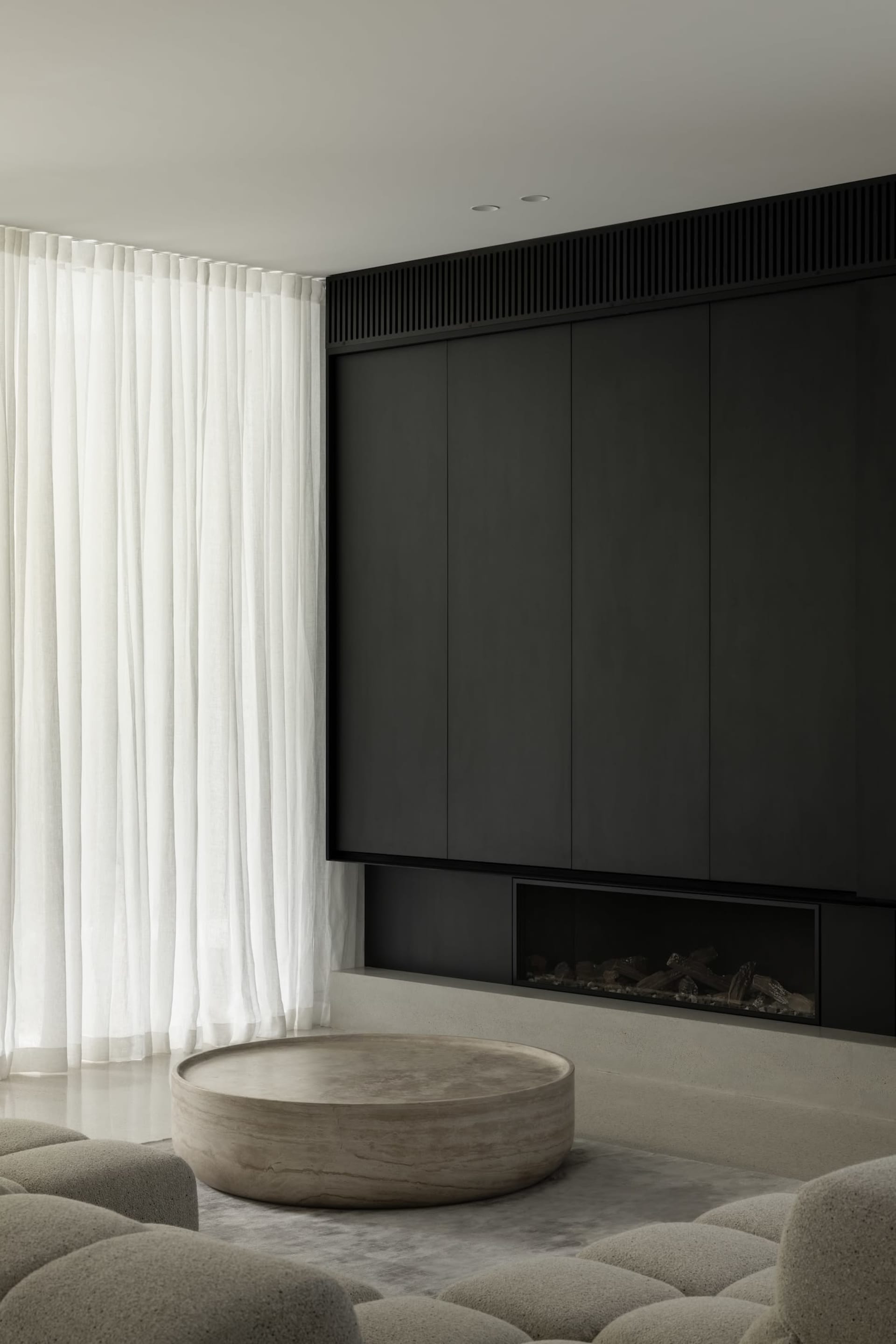 A minimalist living room with white curtains, dark minimalist cabinetry, a circular coffee table, and textured grey seating.