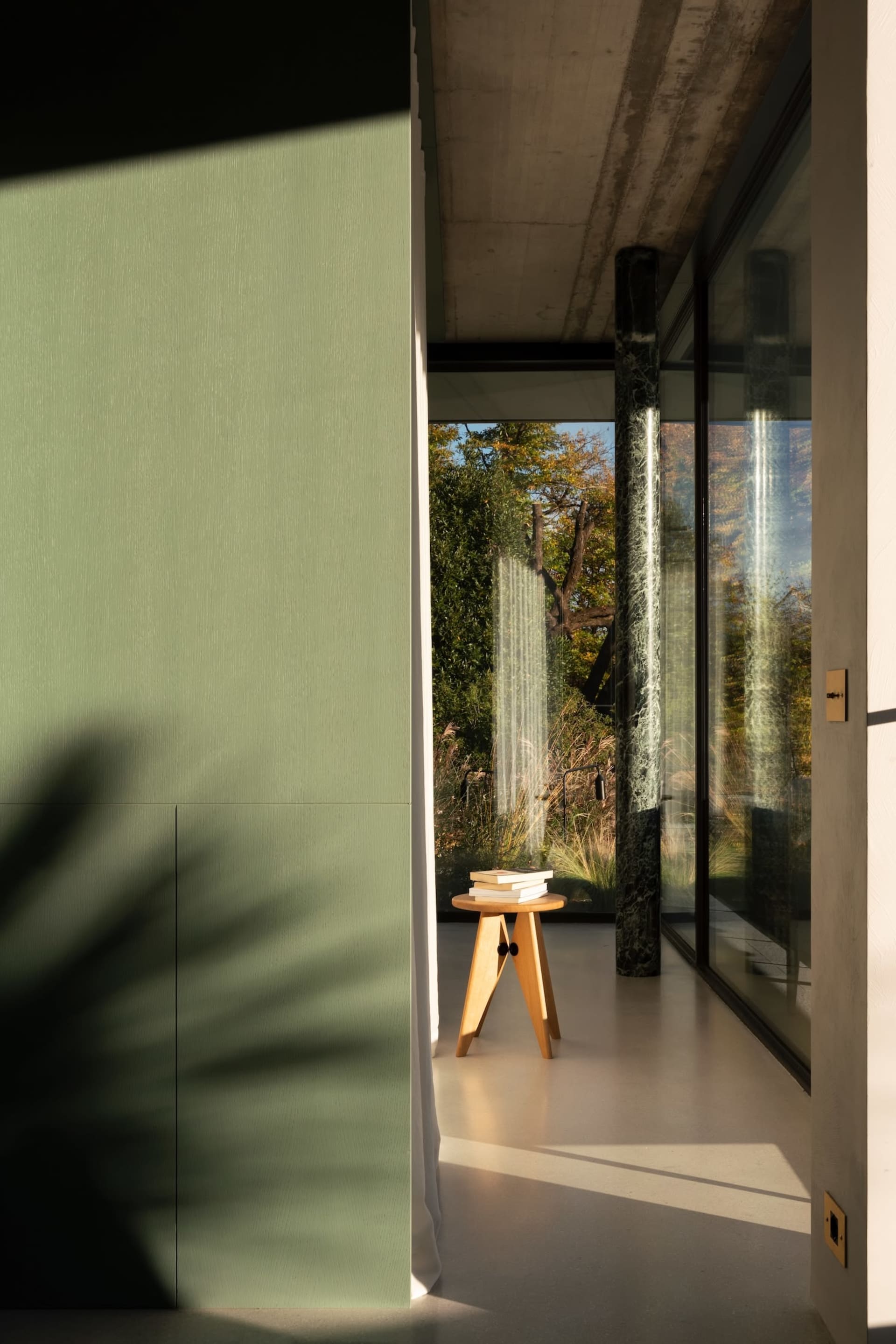 A minimalist interior with concrete walls and glass elements, featuring a small wooden stool with books on it.