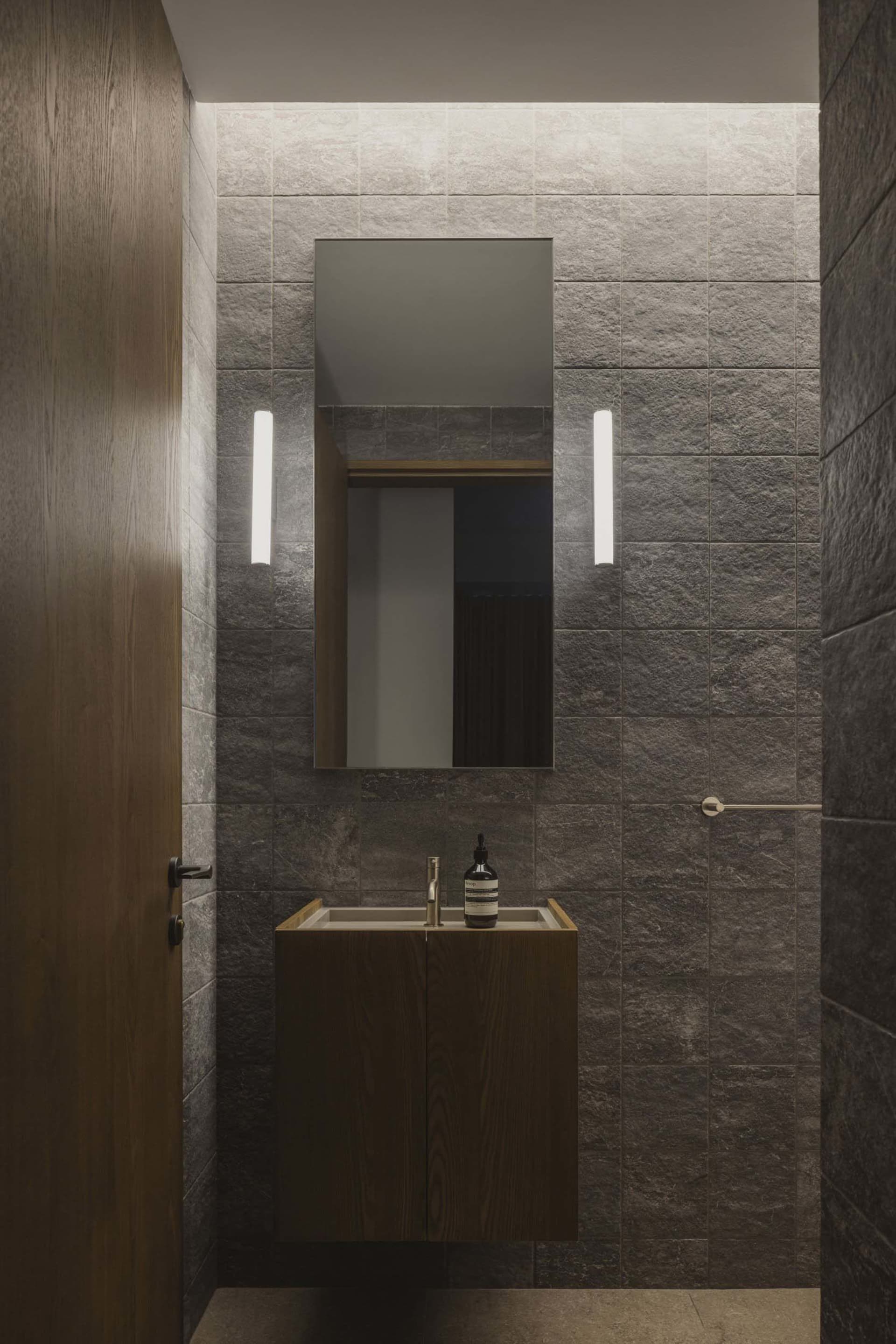 A modern bathroom with stone tiles, a floating vanity, and a rectangular mirror with LED lighting on either side.