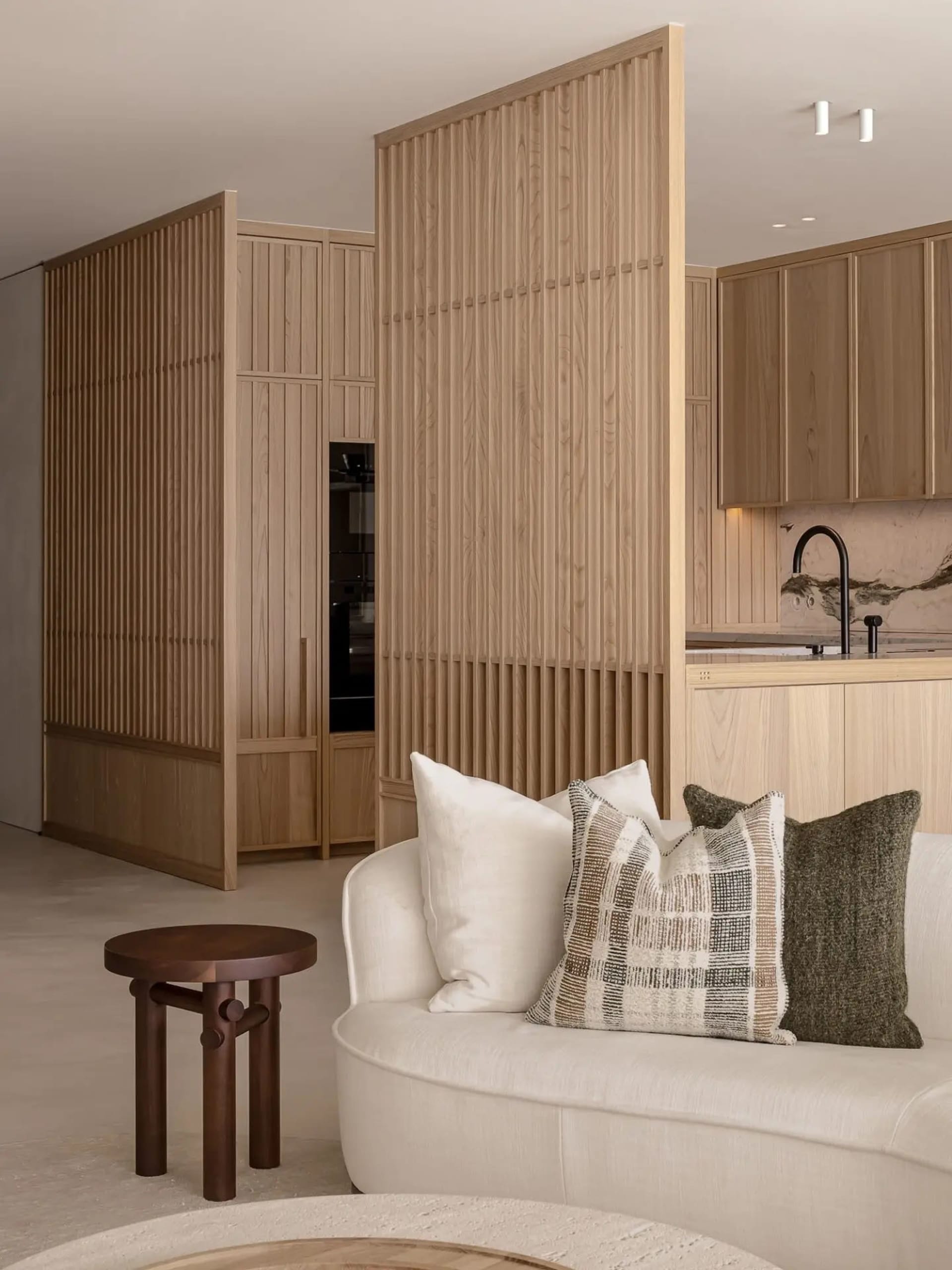 A modern residential interior featuring a white sofa with patterned and solid pillows, wooden furniture, and a kitchen area w