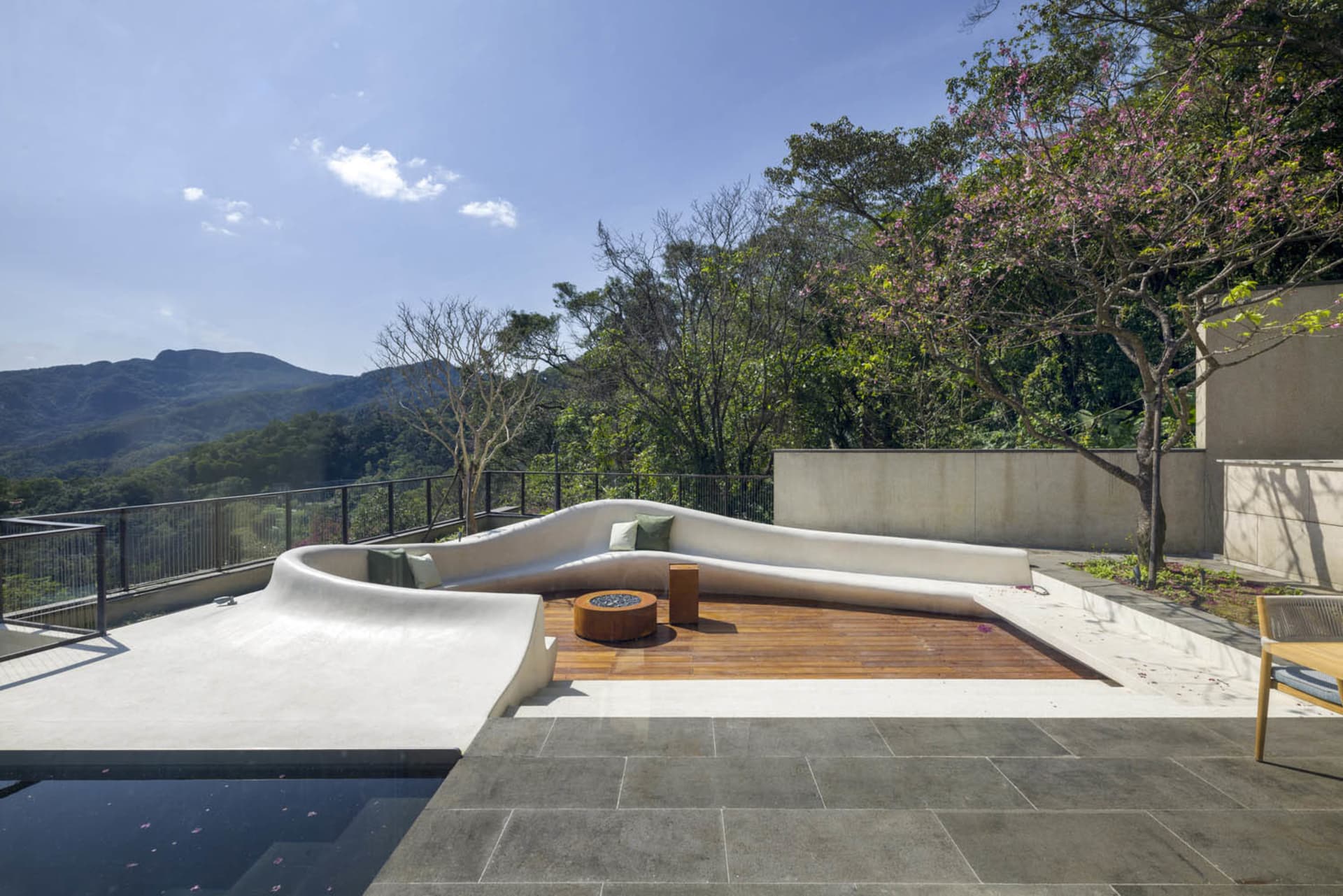 A modern roof deck features a curvilinear concrete seating area with green cushions, a wooden fire pit table, and a view of t